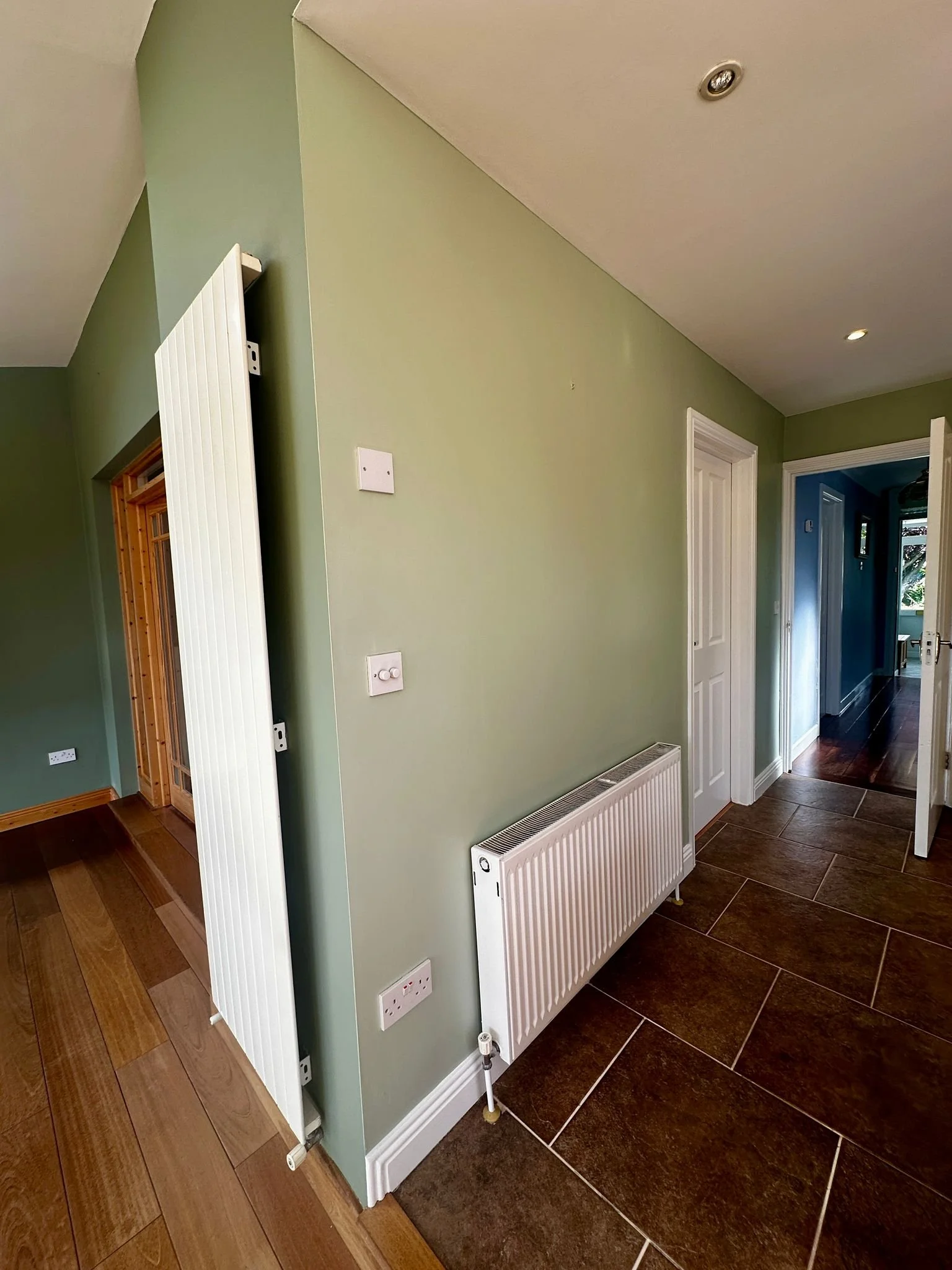 Interior view of a room with a green wall, white radiator, and brown tiled floor. There are three white doors and a partially opened black door leading to a hallway with wooden flooring.