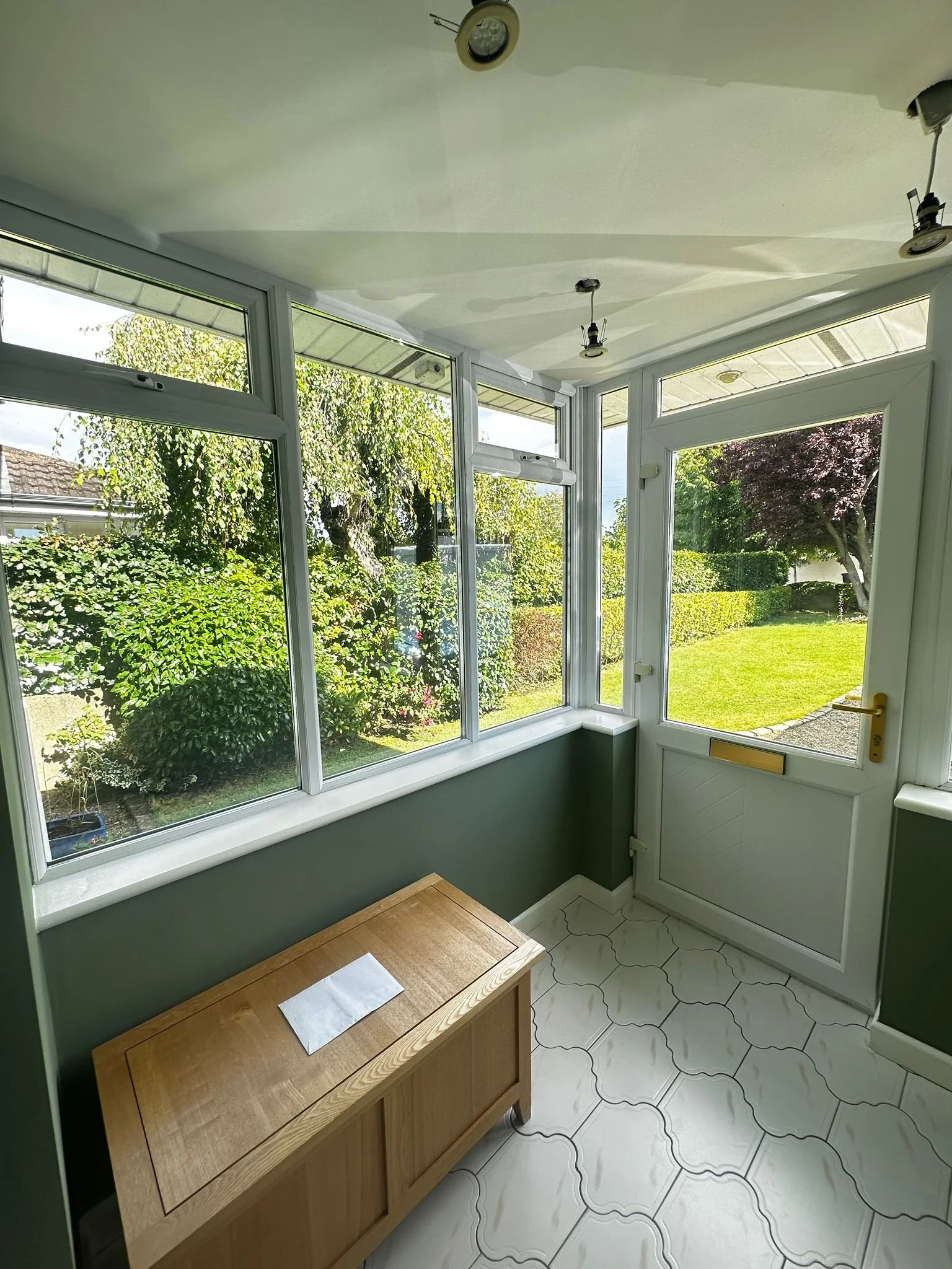 Sunlit enclosed porch with large windows, a wooden chest, and a view of a green garden with trees and shrubs.