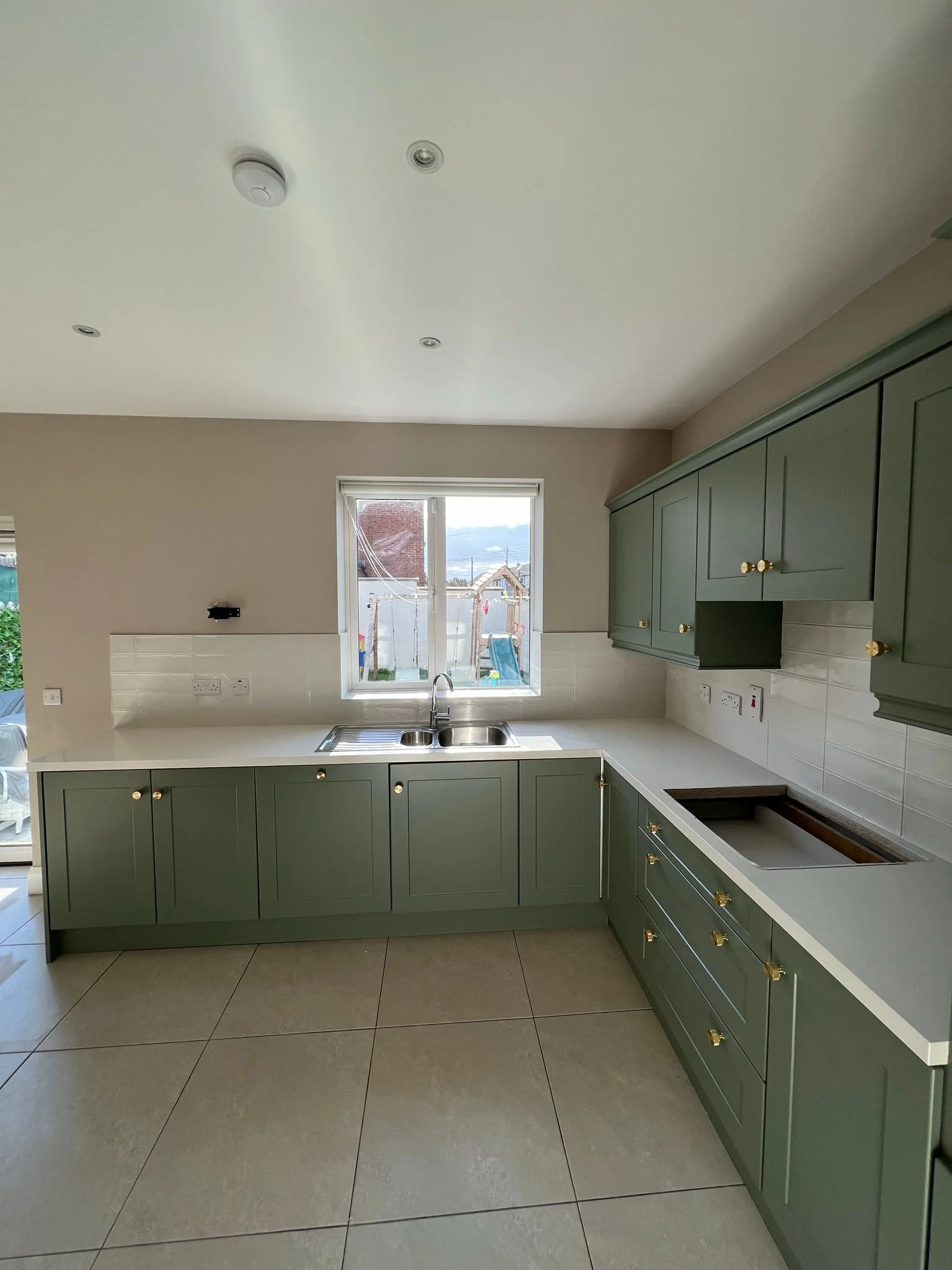 Kitchen with green cabinets, white countertop, sink under window, and tiled backsplash, with an empty space where the stove should be.