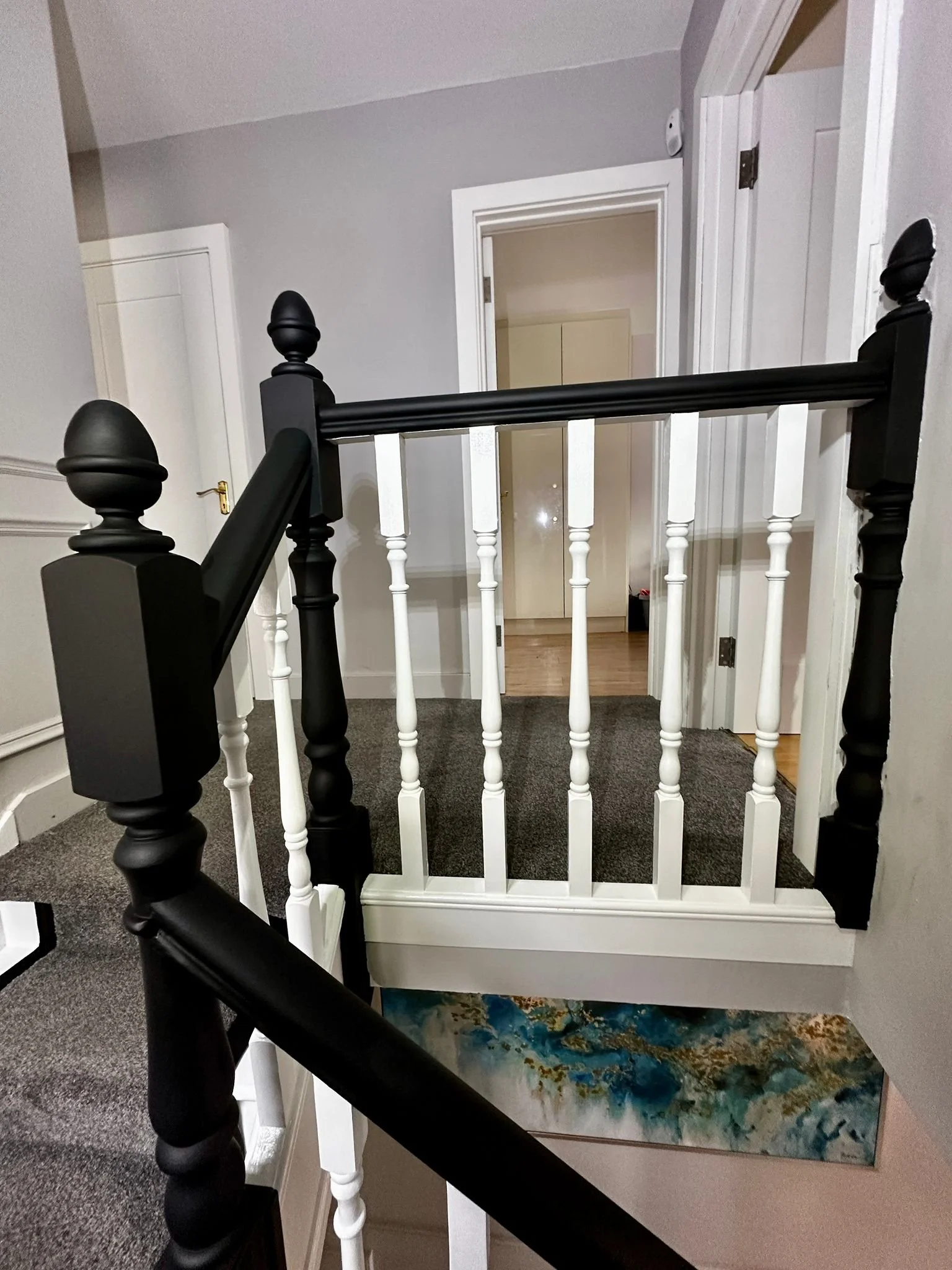 Interior view of a staircase landing with black and white wooden railing, beige carpeted floors, and a door open to a room with beige walls and a closed door.