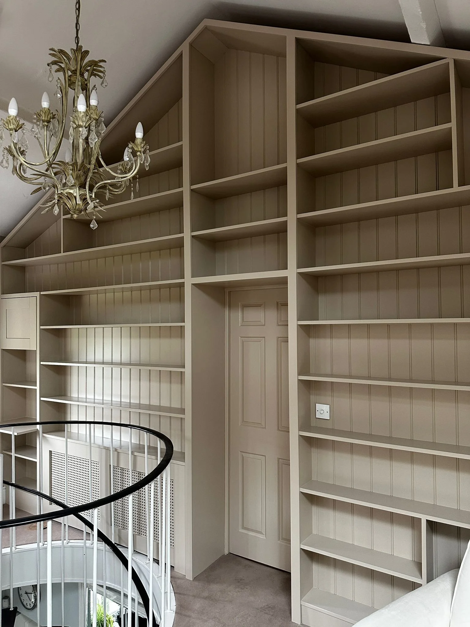 Empty beige built-in bookshelf with multiple shelves near a staircase and a chandelier in a room with sloped ceiling.
