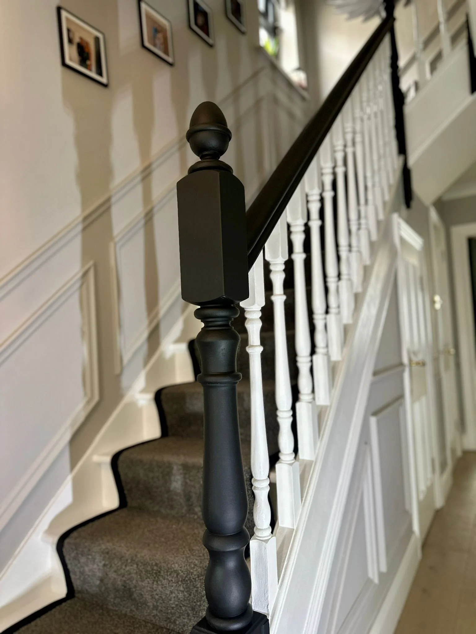 Close-up of a staircase handrail with white spindles, black cap and top, with a gray carpeted stair below, in a hallway with framed photos on the wall.