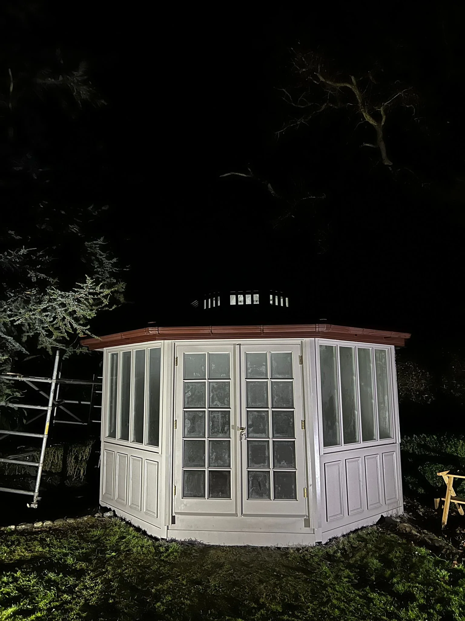 A white, octagonal sunroom with glass panes on the doors and windows, illuminated at night with surrounding trees and grass.