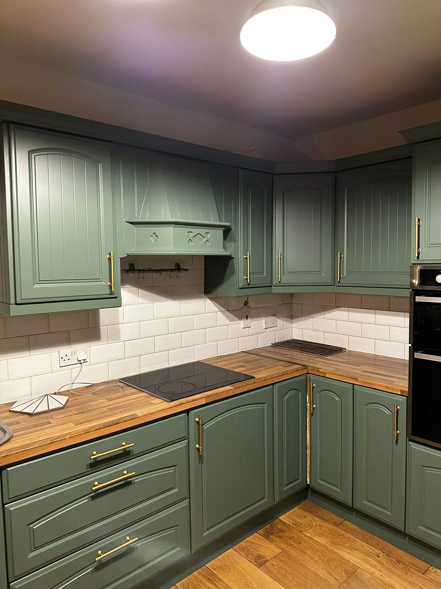Green kitchen cabinets with gold handles, wooden countertop, white brick tile backsplash, electric stovetop, and ceiling light fixture.