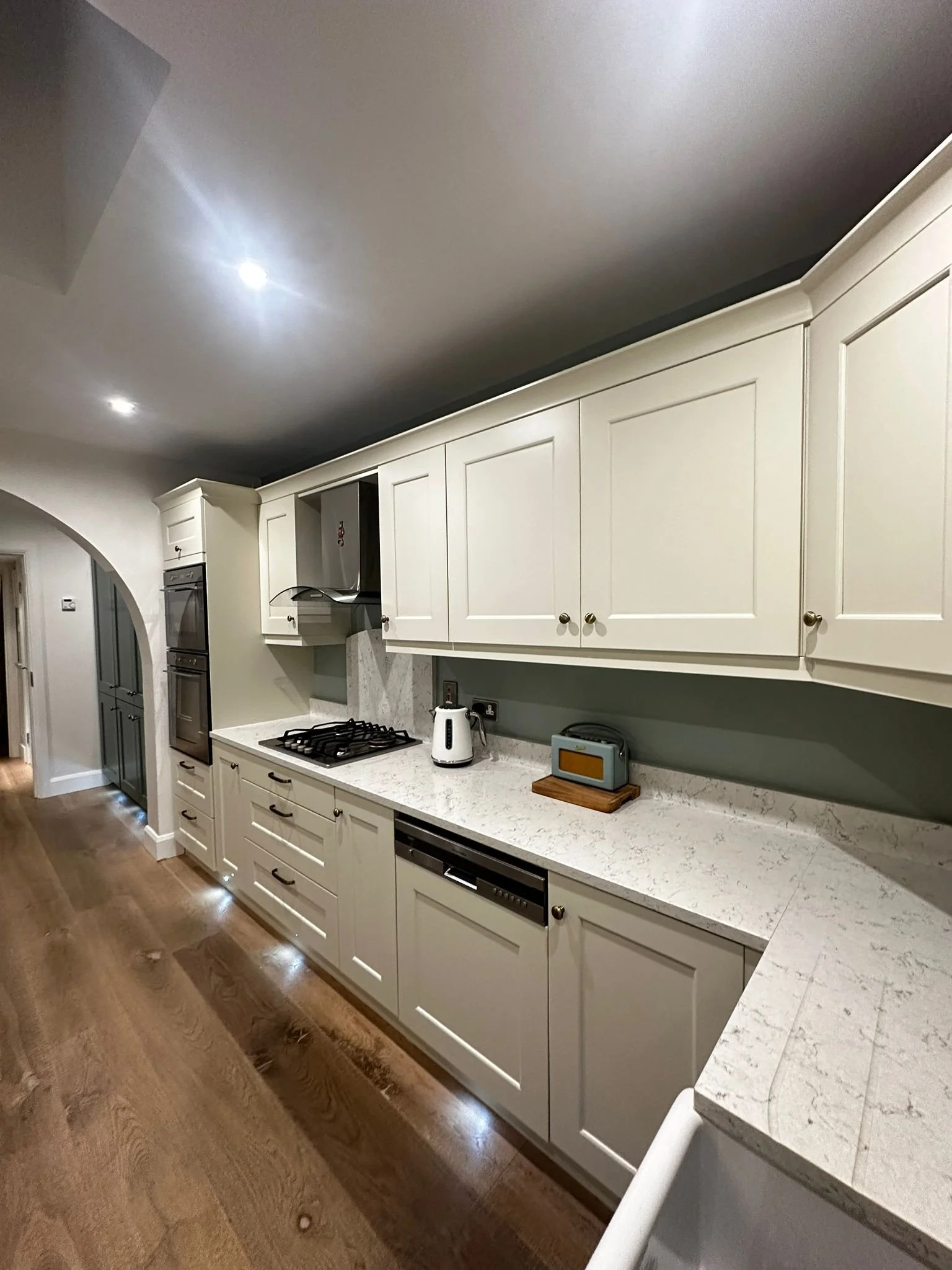 Modern kitchen with white cabinets, marble countertops, two built-in ovens, a cooktop, a range hood, a toaster, and a wooden cutting board on the counter.