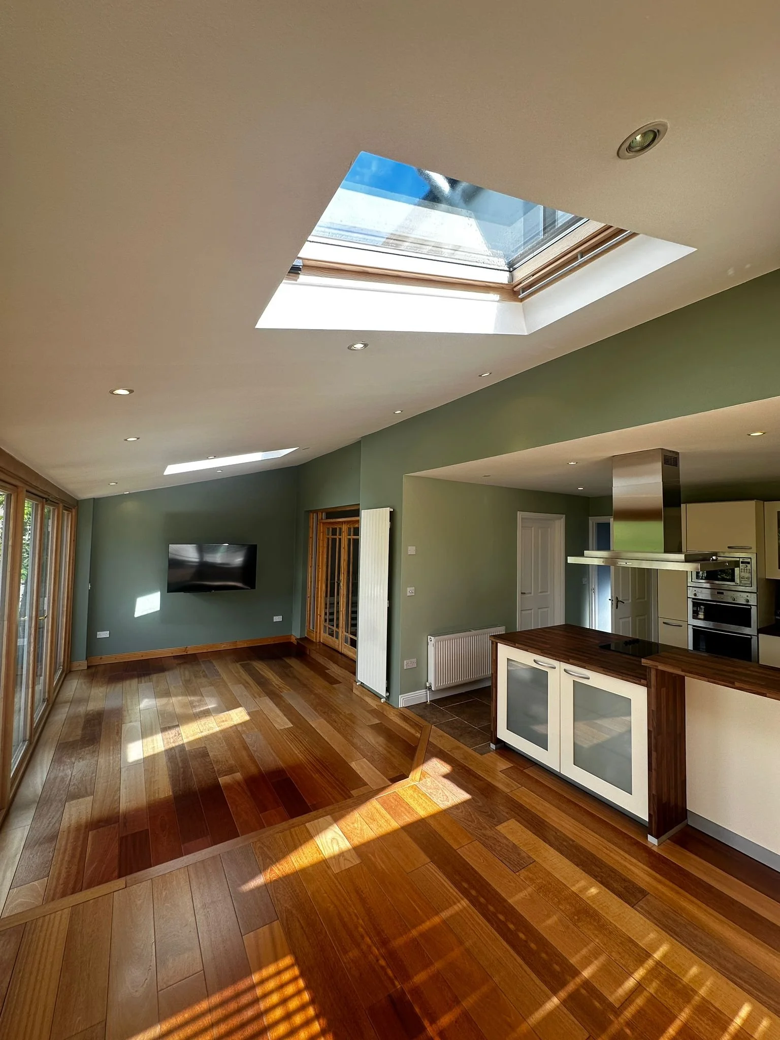 Sunlit open living space with hardwood floors, a wall-mounted TV, a skylight in the ceiling, and a kitchen area with an island and modern appliances.