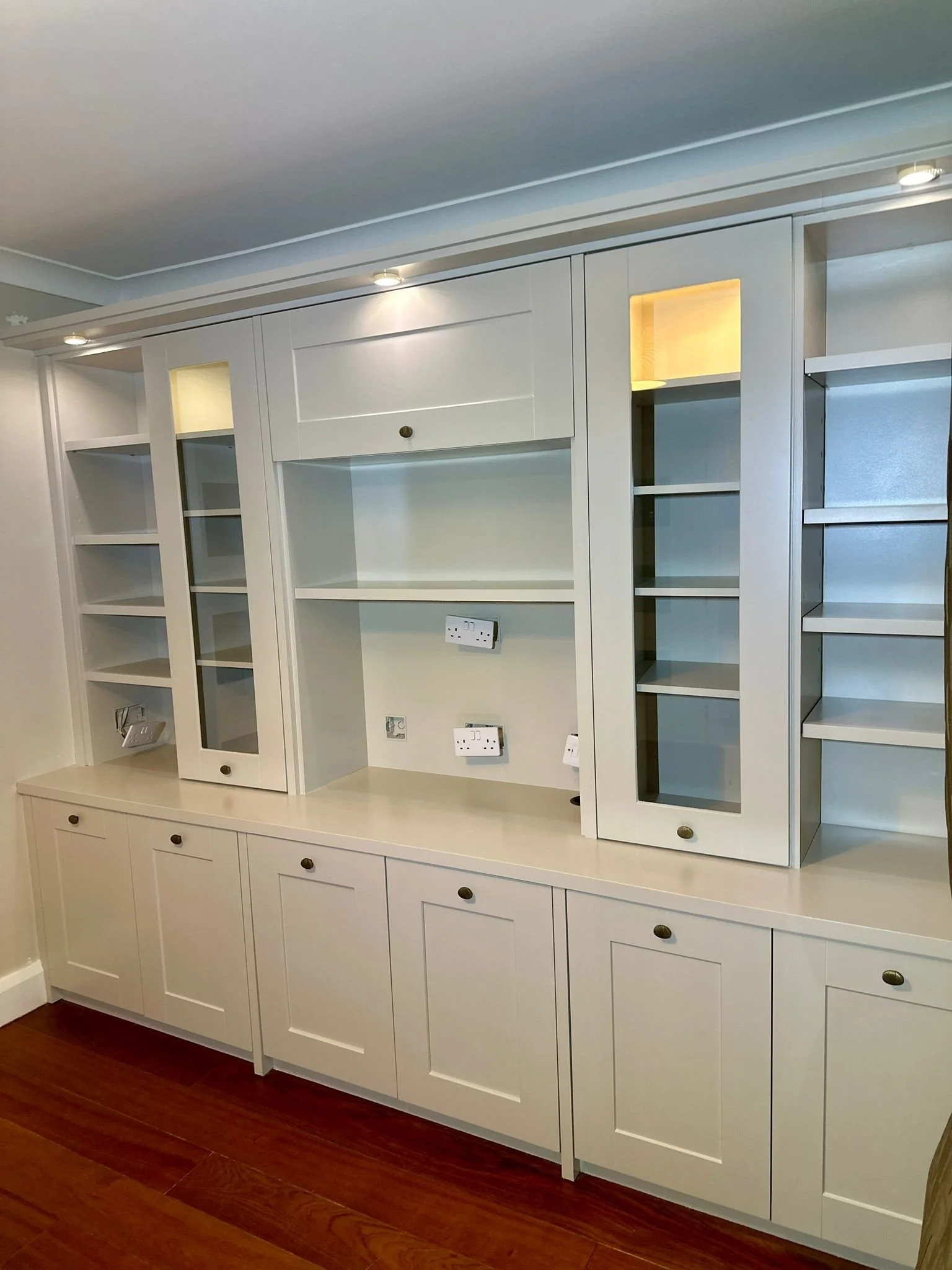 White built-in cabinet with shelves, glass doors, and electrical outlets on the wall underneath. Wooden flooring.