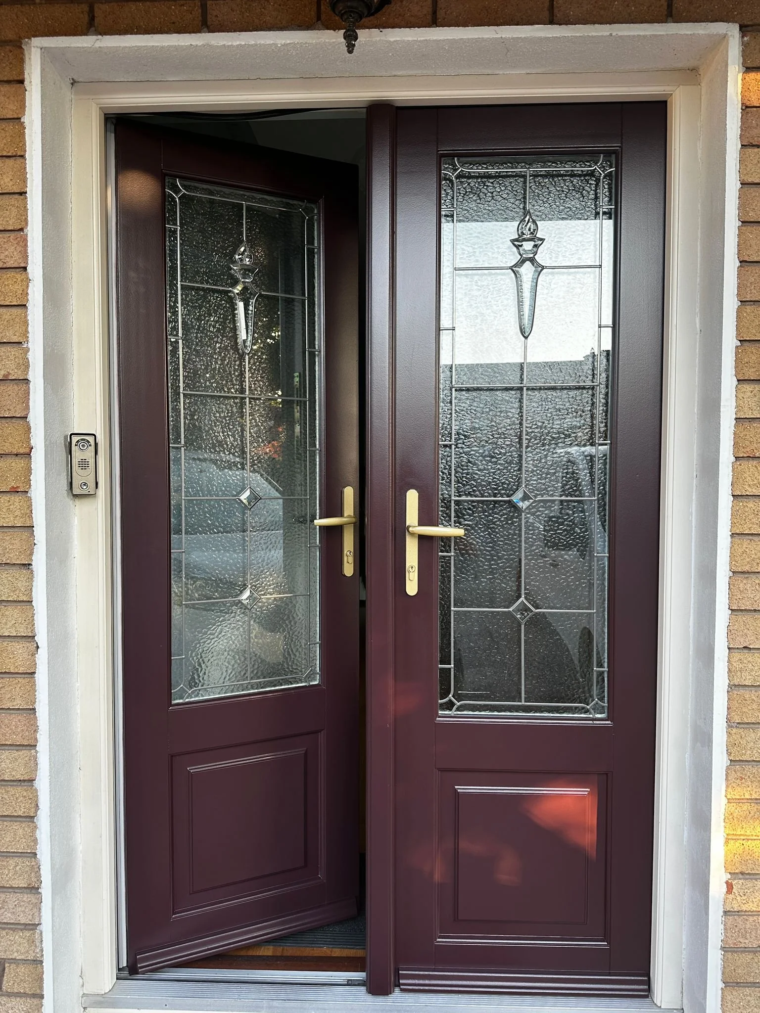 Front door with two glass panels with decorative patterns and a brass handle, set in a brick house with a white frame.