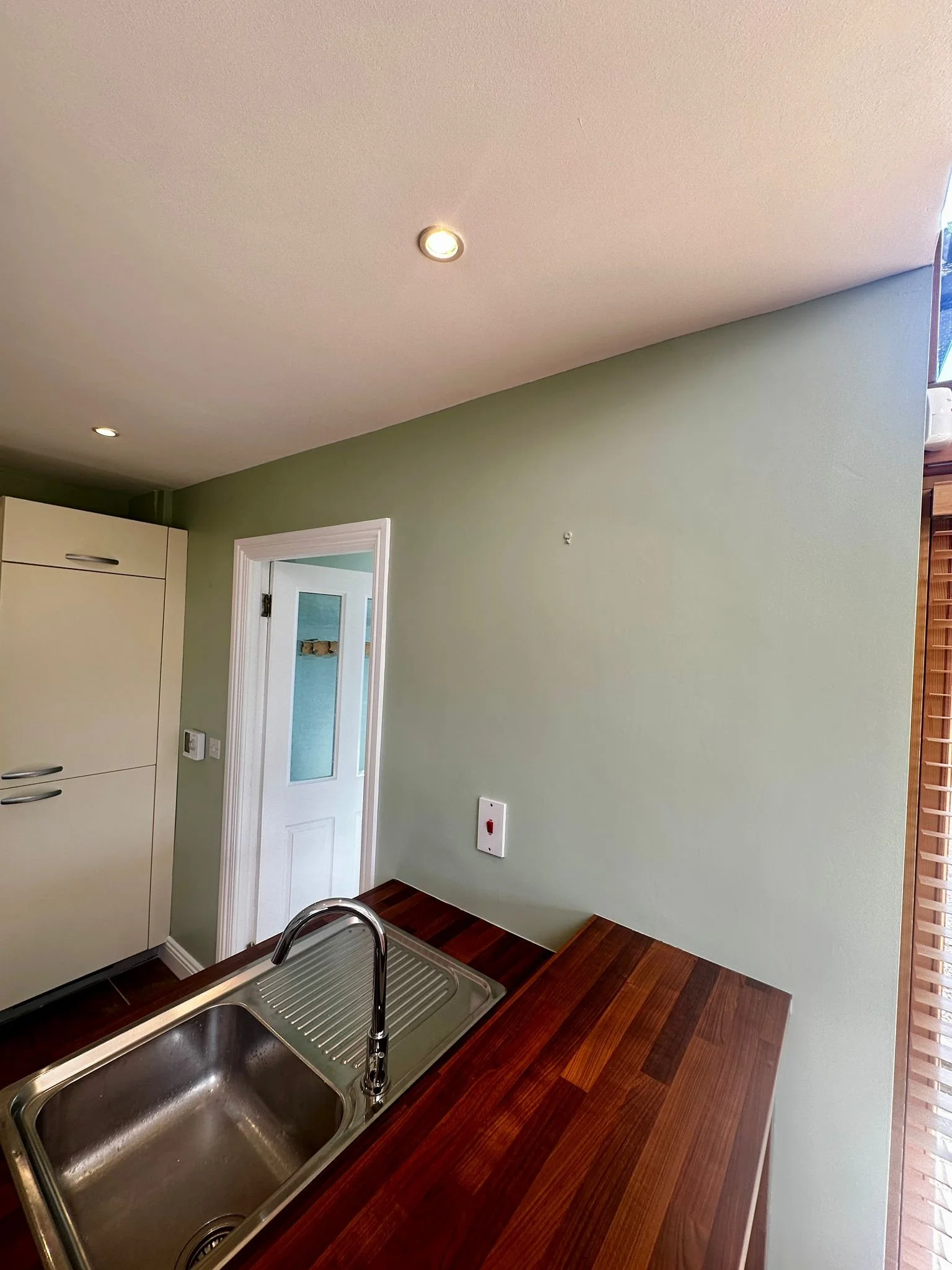 Kitchen with a stainless steel sink, wooden countertop, and a green wall with a small hole and a thermostat. Part of a white door and window with wooden blinds are visible.