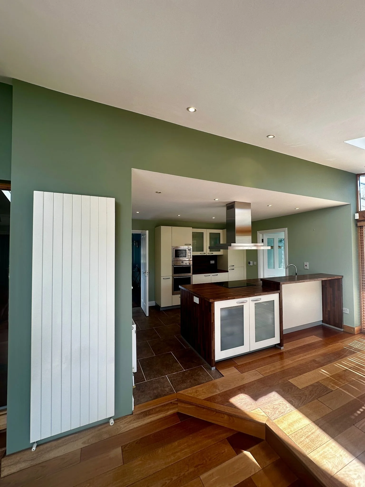 Modern kitchen with green walls, wooden floors, and white cabinets, featuring a central island and stainless steel appliances.
