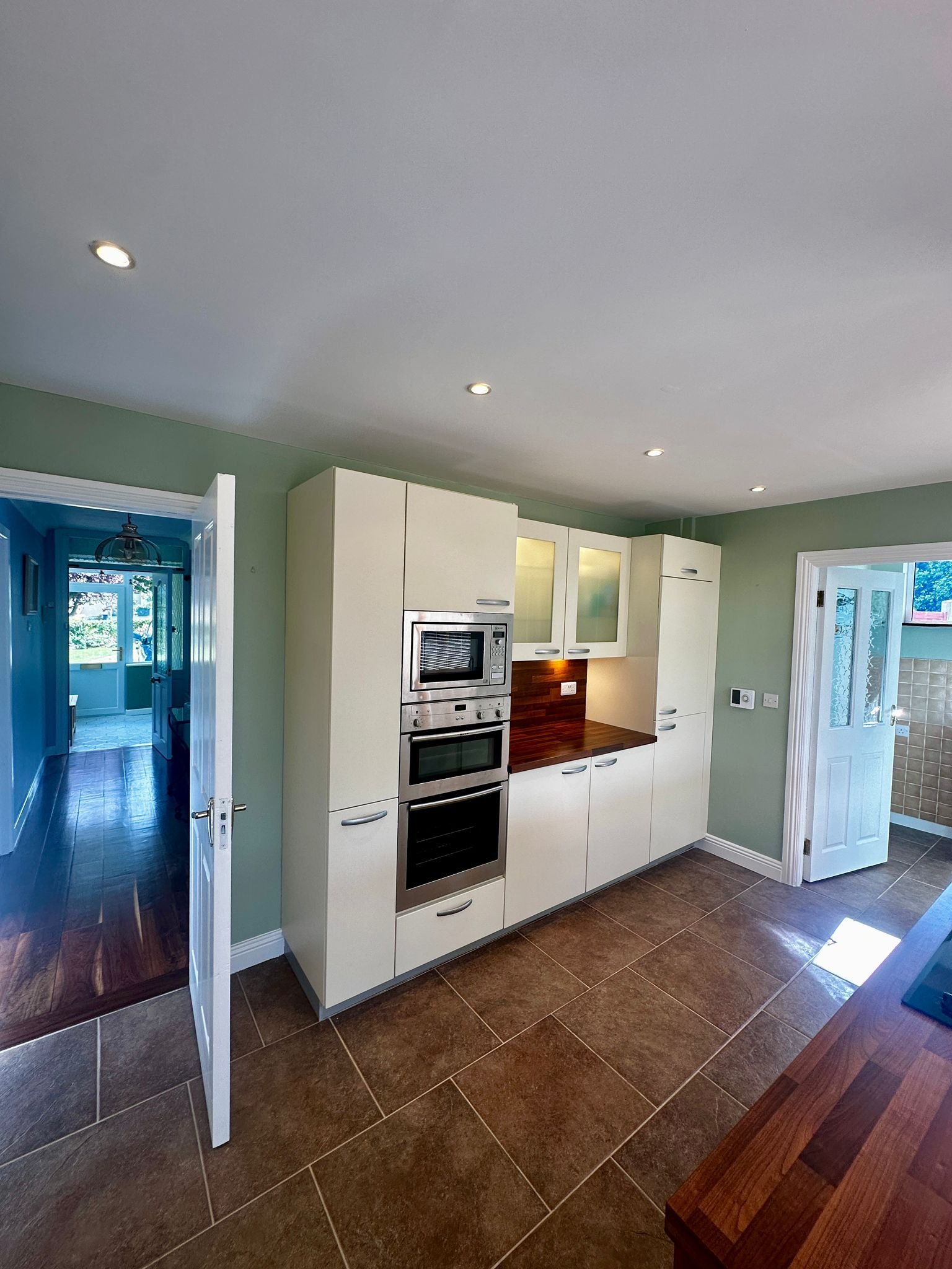 Kitchen with built-in oven and microwave, white cabinets, wooden countertop, green walls, brown tiled floor, open door to hallway and adjacent room with tiles and window.