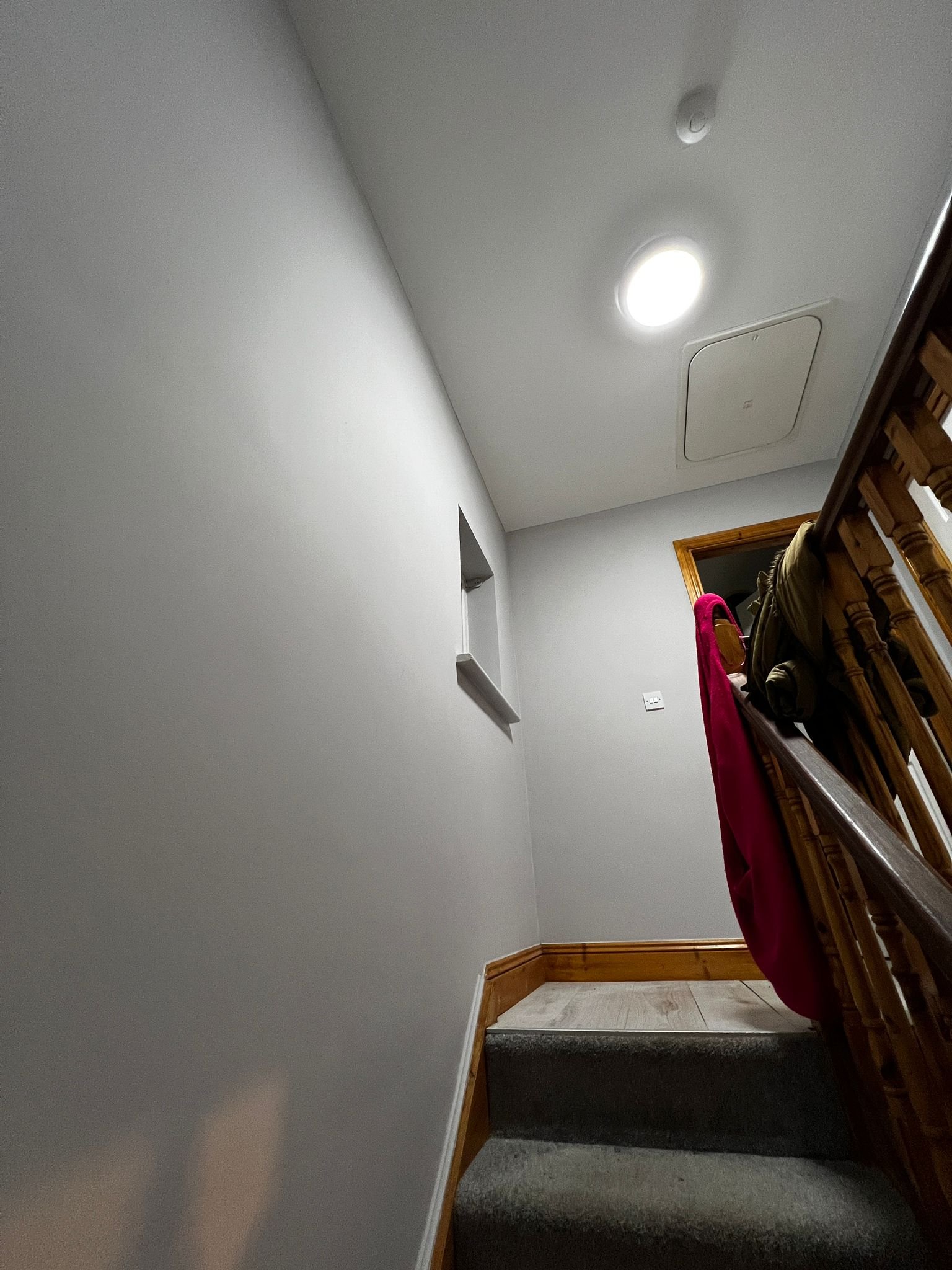 Interior view of a staircase landing with carpeted steps, a white wall, a small wall niche, wooden trim, a ceiling light, smoke alarm, and a door opening to another room. Clothes hang over the staircase railing.