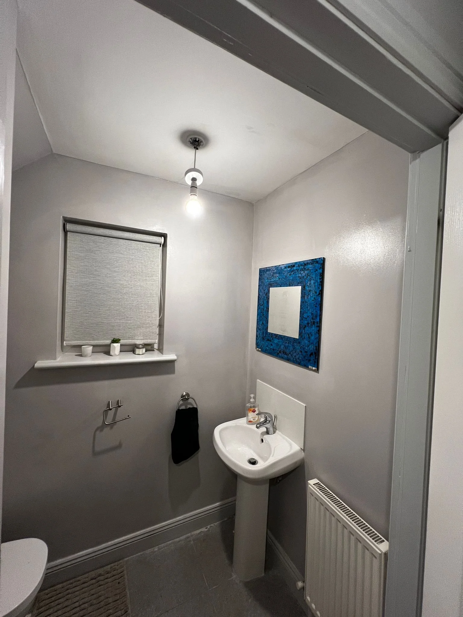 A small bathroom with white walls, a pedestal sink, a black hand towel, a window with a roller blind, a mirror with a blue frame, a radiator, a ceiling light, and a soap dispenser on the sink.