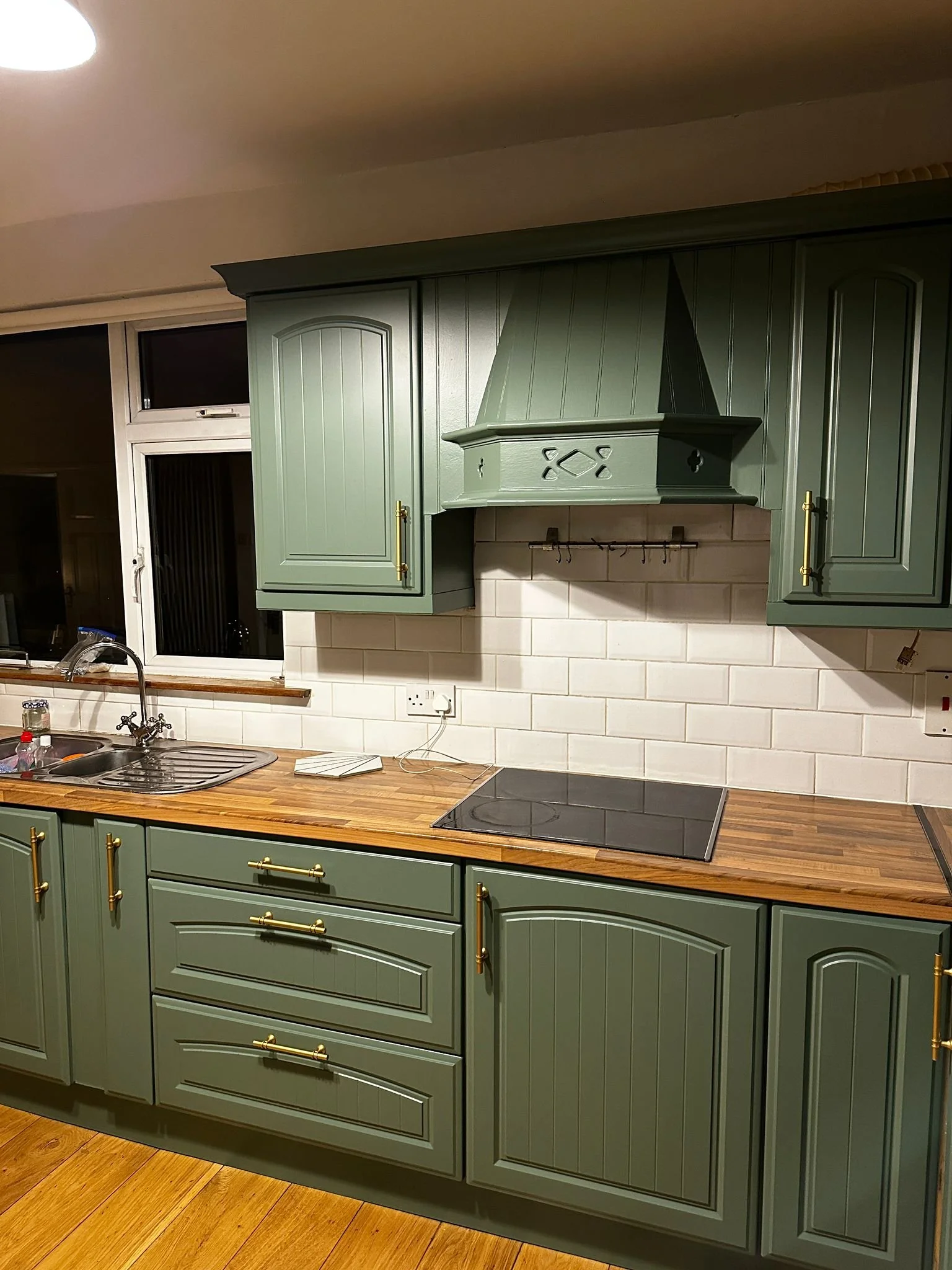 Green kitchen cabinets with brass handles, a wooden countertop, a double sink, and a black electric cooktop. There is a window above the sink, white tiled backsplash, and a range hood.