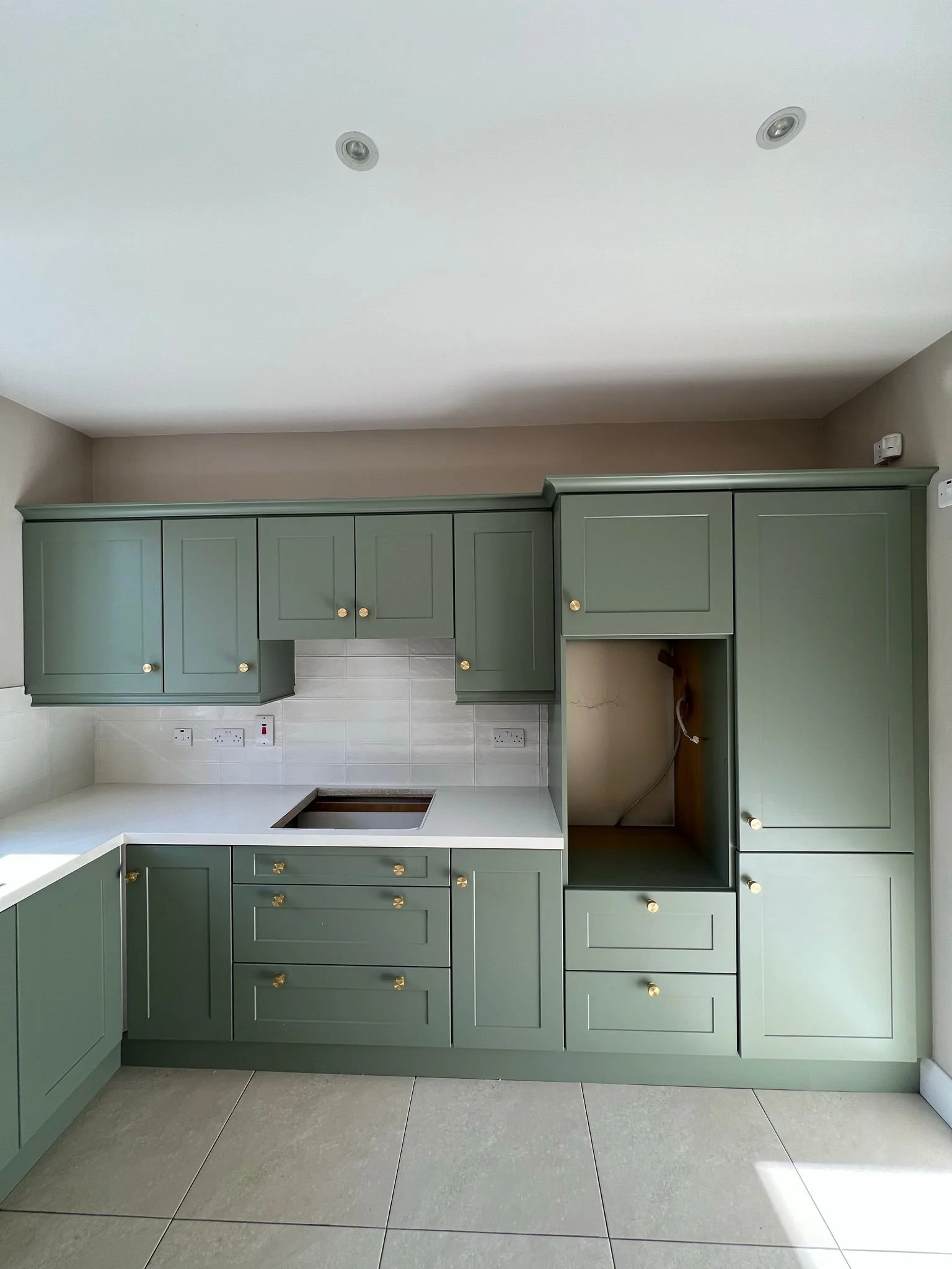 Green kitchen cabinets with gold knobs, a white countertop, and a partially open space where a stove would be installed. Two electrical outlets are visible on the backsplash, and the kitchen has beige tiled flooring.