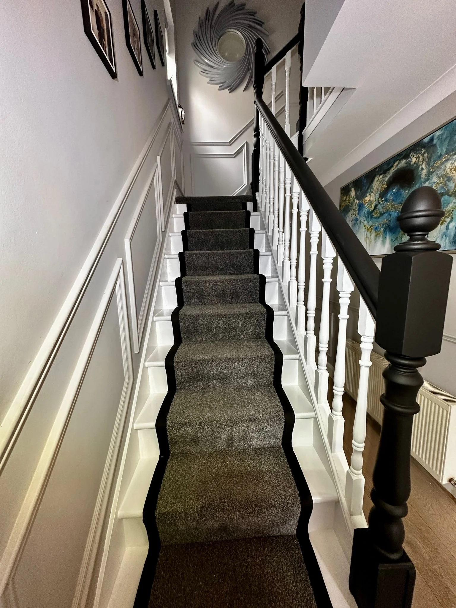 A staircase with a black and gray runner rug, white railing with black handrail, beige walls, and artwork on the wall.