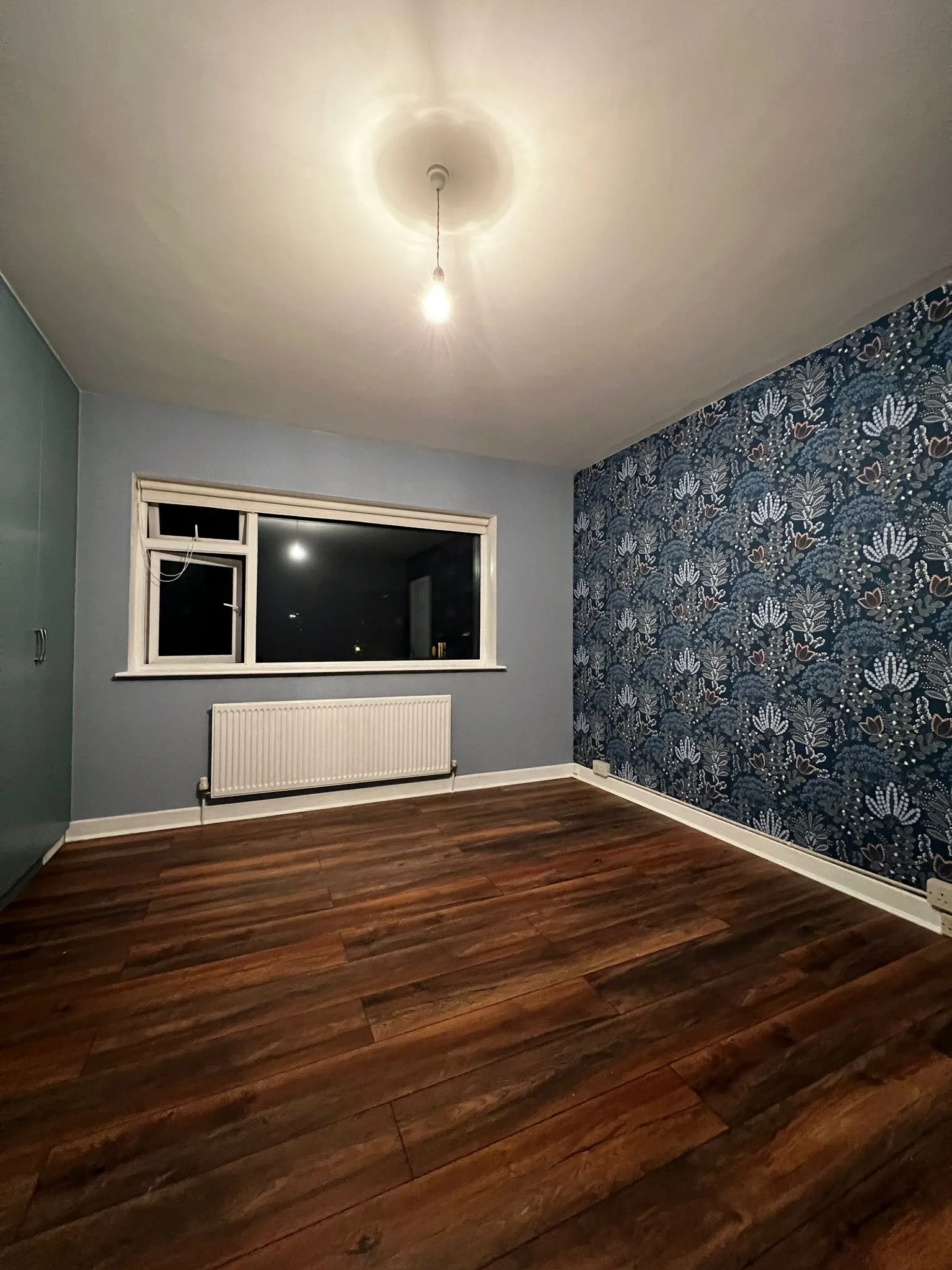 Empty room with a window, radiator, wooden floor, and a wall with floral wallpaper.