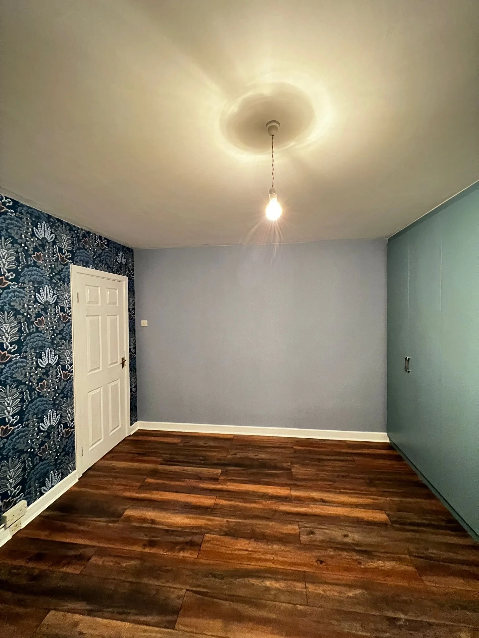Empty room with wooden flooring, a decorated wallpapered wall on the left, a plain wall in the middle, and a large built-in wardrobe on the right. A single lightbulb hanging from the ceiling is turned on.