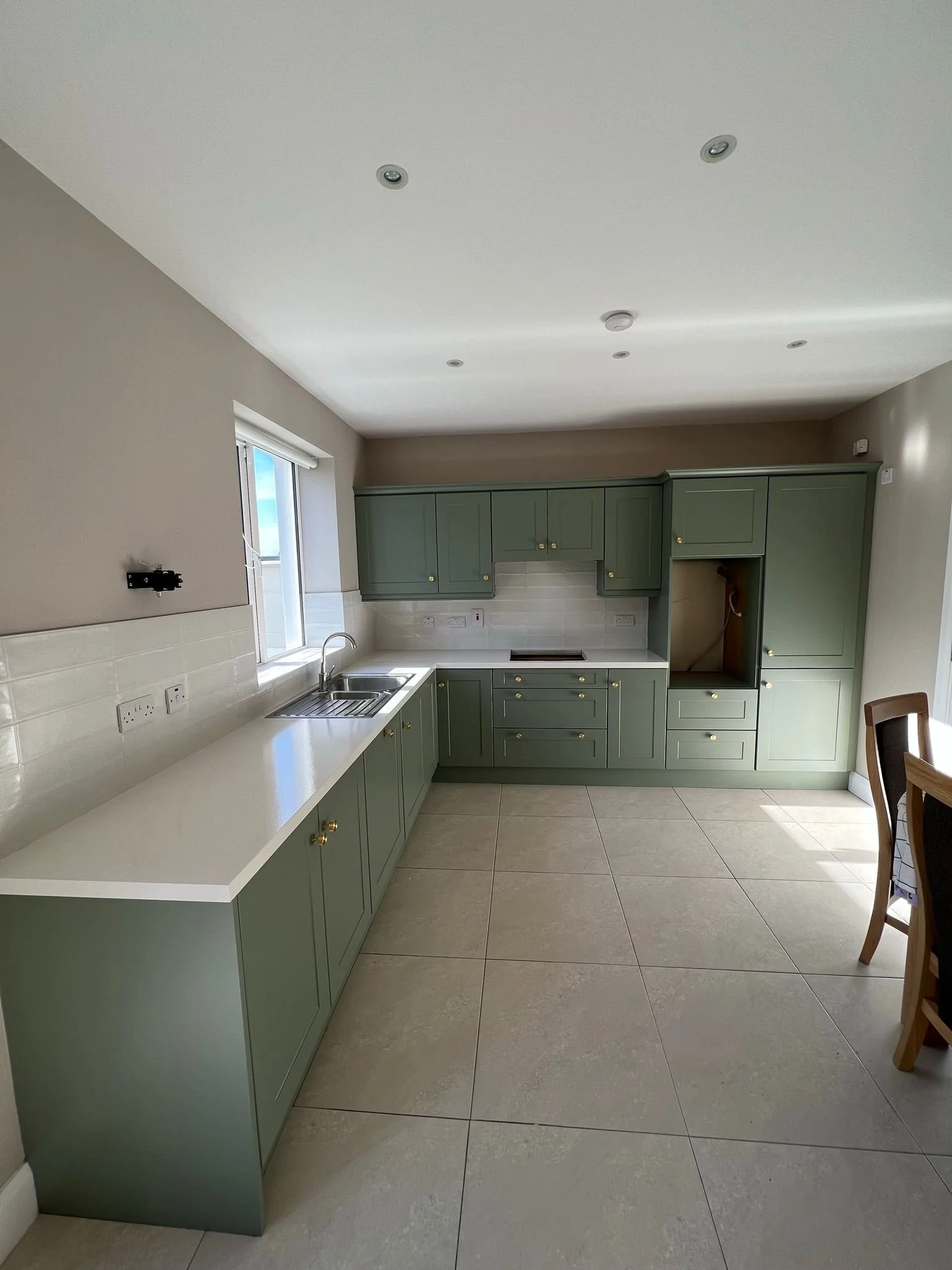 Empty kitchen with green cabinets, white countertops, a sink by a window, light-colored tile floor, and some electrical outlets.