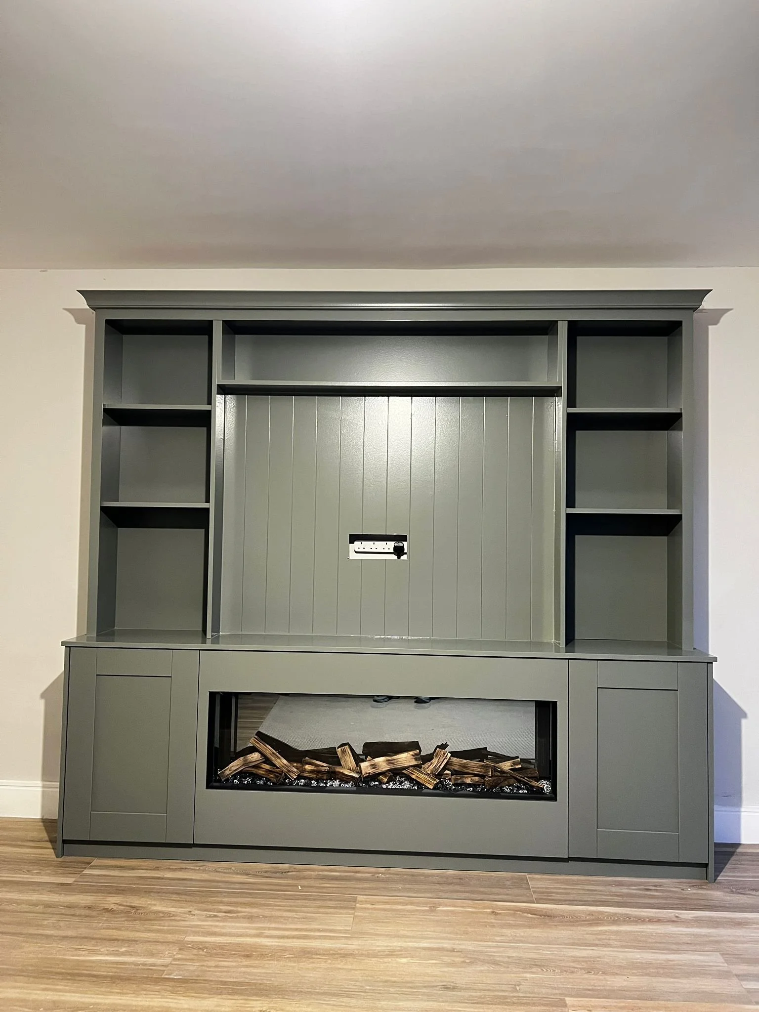 Gray built-in fireplace and bookshelf unit with open shelves, a central section with a mount for a TV, and a glass-enclosed simulated fireplace with firewood inside, against a white wall with wood flooring.