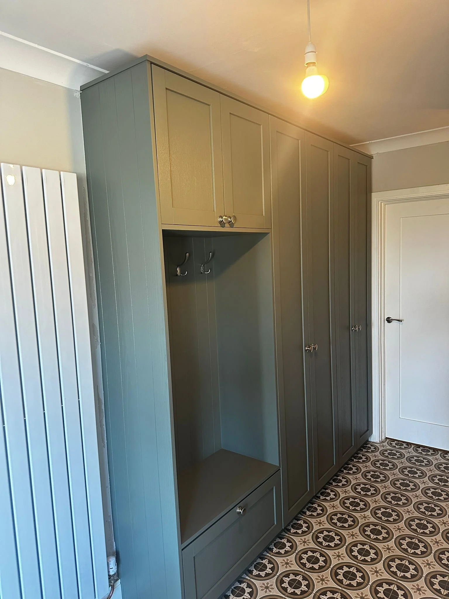 Green built-in wardrobe with silver knobs, hooks, and a small drawer at the bottom, located in a room with patterned black and white tile flooring, a white door, a white radiator, and an exposed lightbulb hanging from the ceiling.