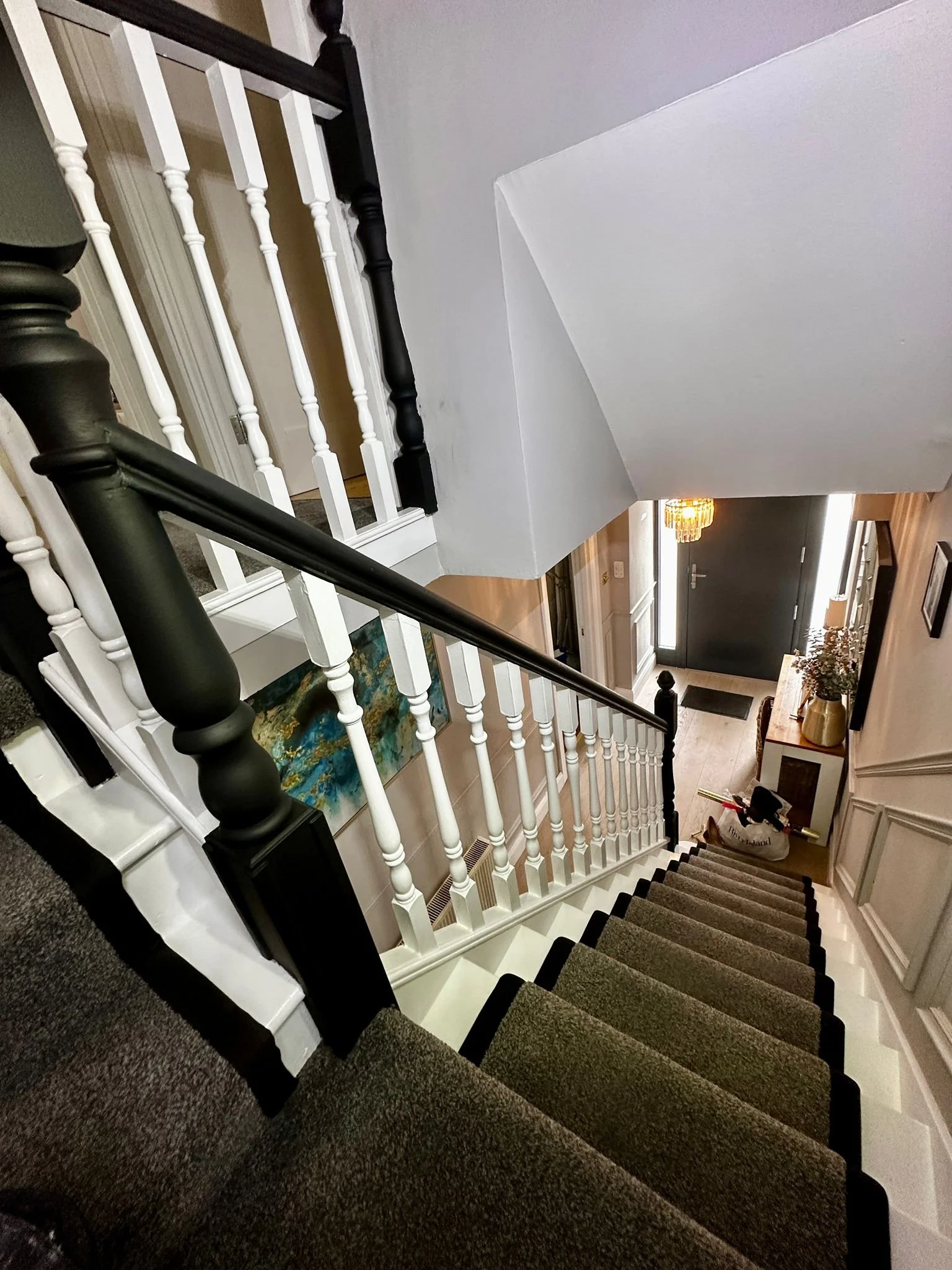 Viewing down a carpeted staircase with black and white railings, leading to a hallway with a black front door, a chandelier, a coat rack, and a side table with a vase of flowers.