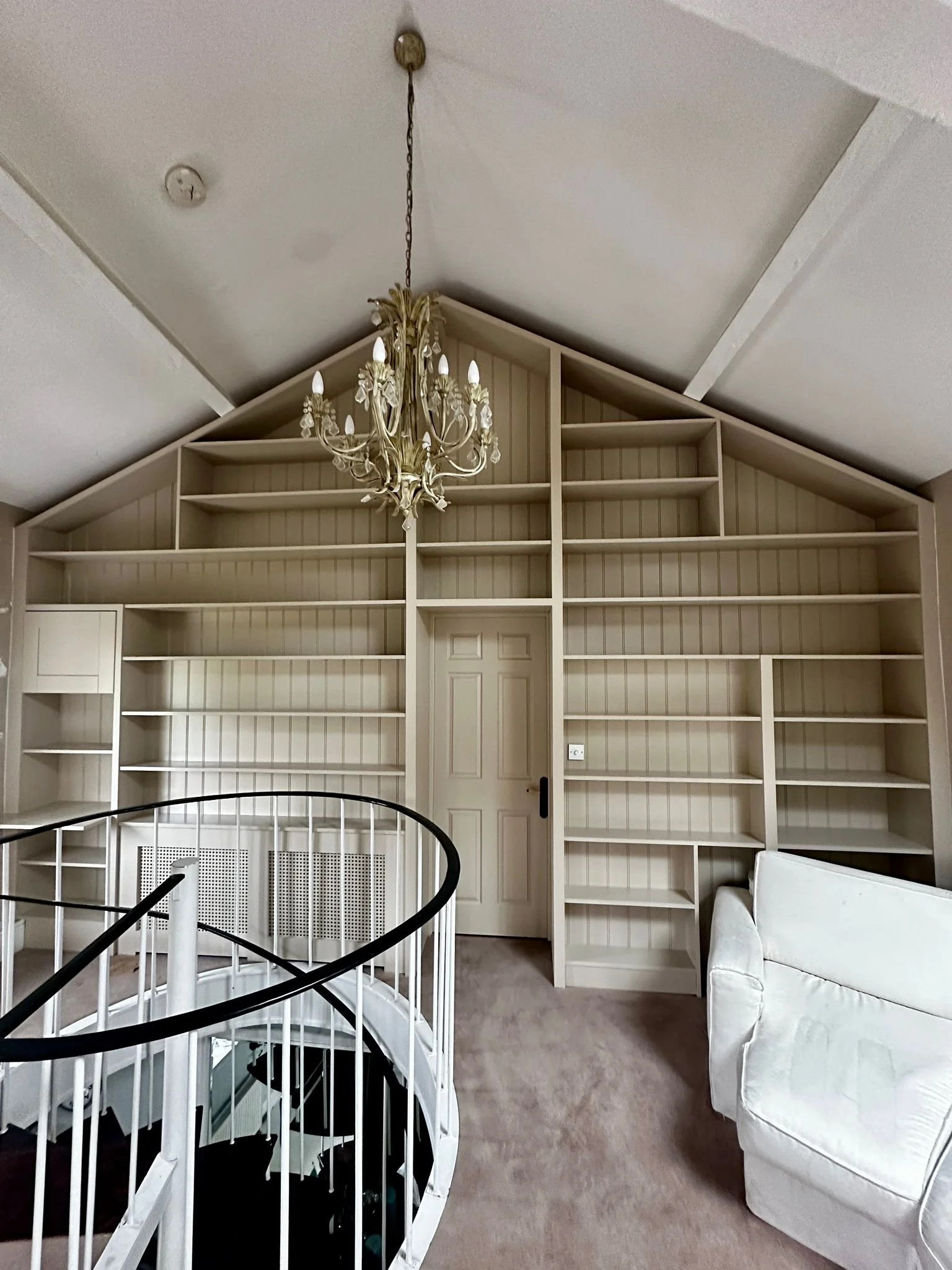 Empty built-in bookshelf with multiple white shelves, a closed corner door, a chandelier hanging from a sloped ceiling, a white couch, and a staircase with a black handrail in a room with beige carpet and walls.