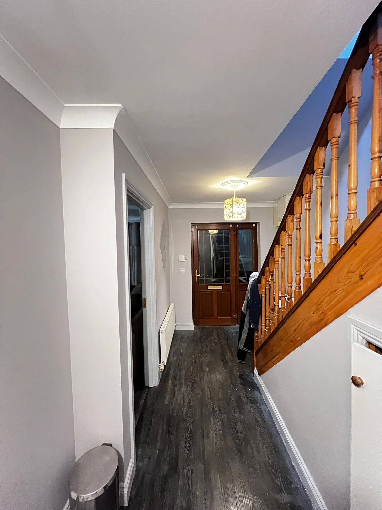 Inside view of a house entrance with a dark door, stairs with wooden railing, coat hanging, and a ceiling light.