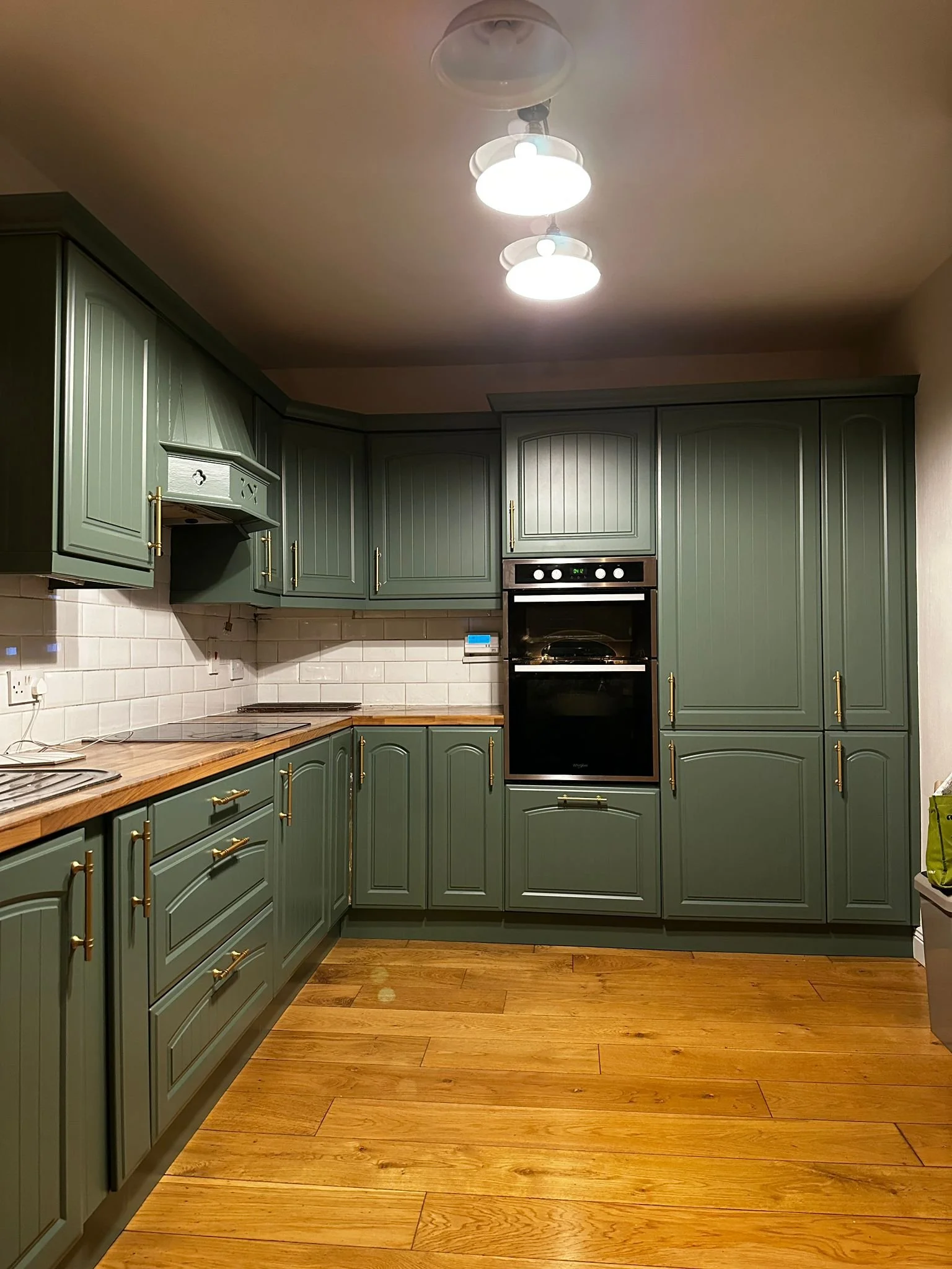 Green kitchen cabinets with gold handles, a wooden countertop, a built-in black oven, white subway tile backsplash, and wooden flooring, illuminated by ceiling lights.