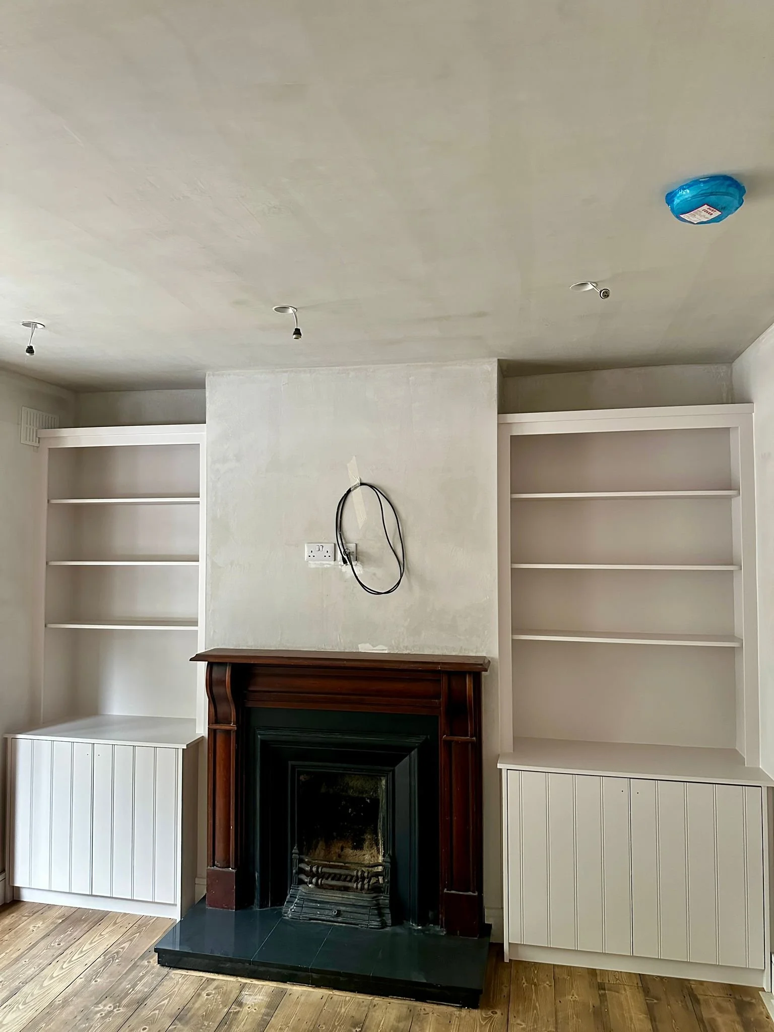Interior of a room with a fireplace, two built-in bookshelves on either side, and unfinished ceiling with exposed light fixtures and a smoke detector.
