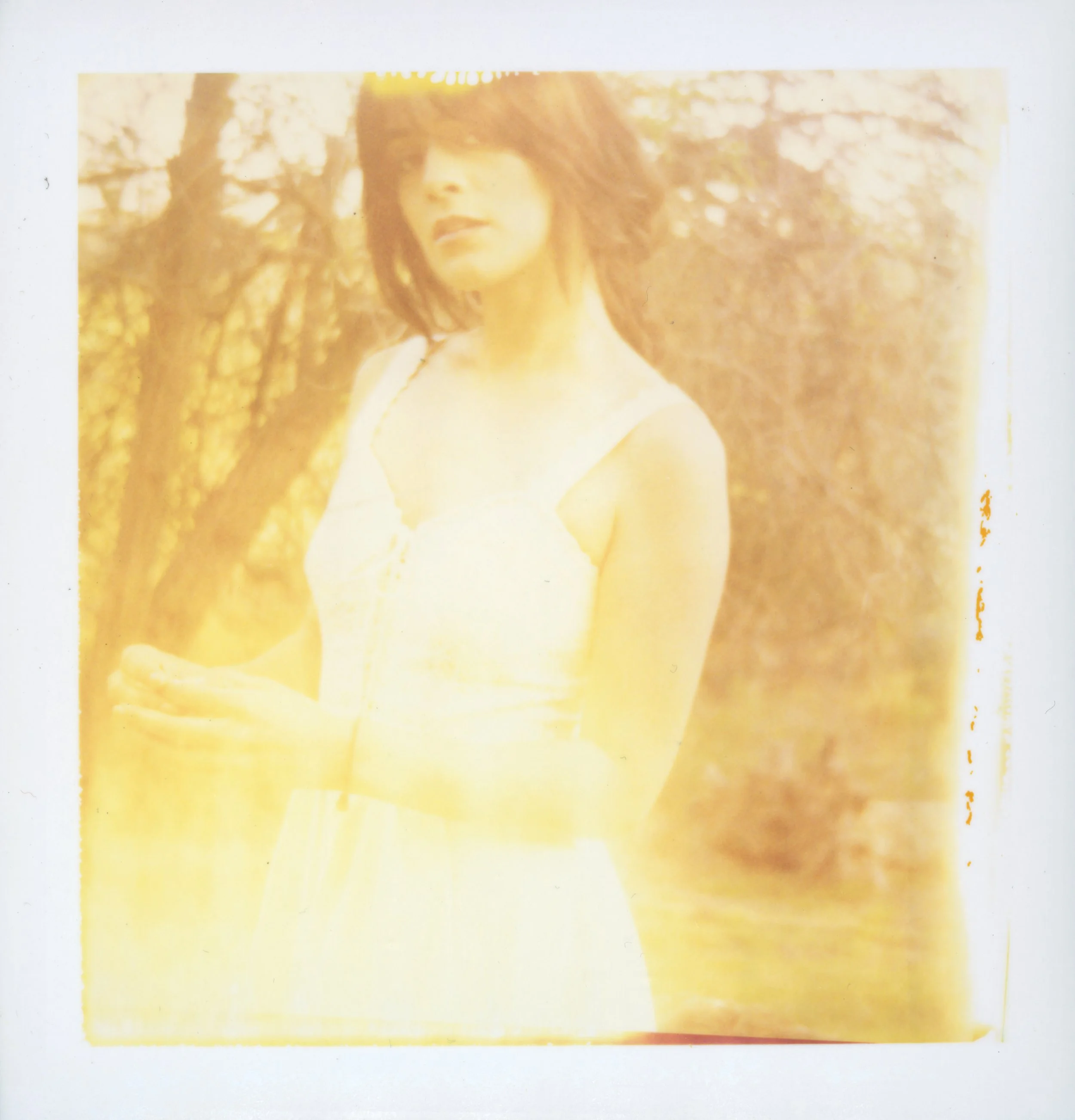A woman with shoulder-length dark hair wearing a yellow dress standing outdoors with trees in the background.