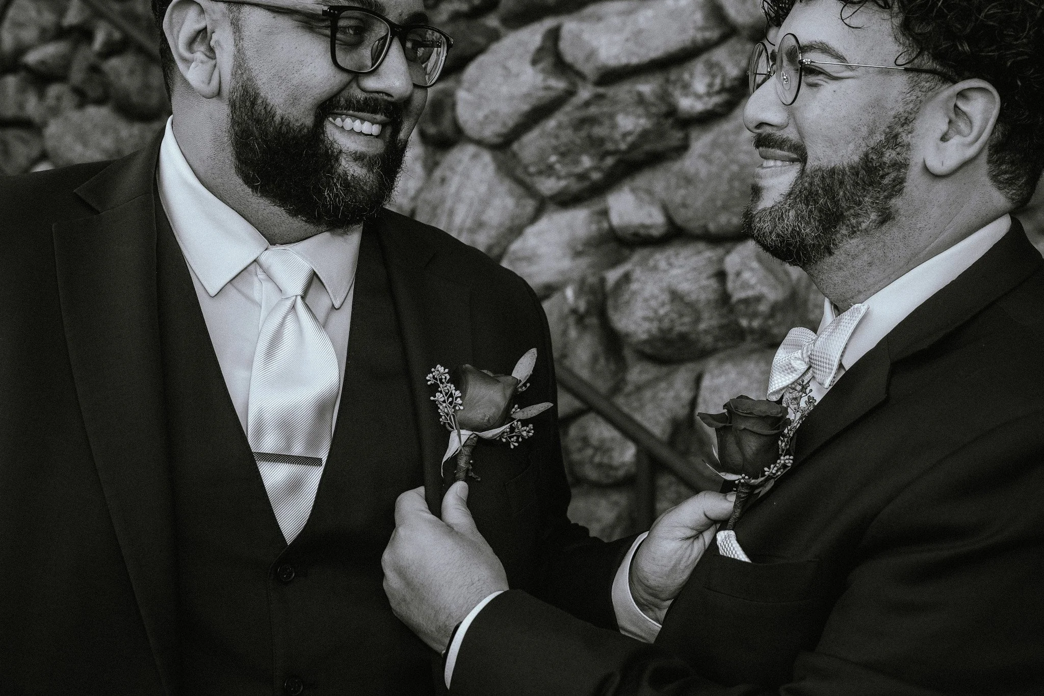 Two men dressed in tuxedos with boutonnieres are smiling and looking at each other. One man is holding the other's lapel.