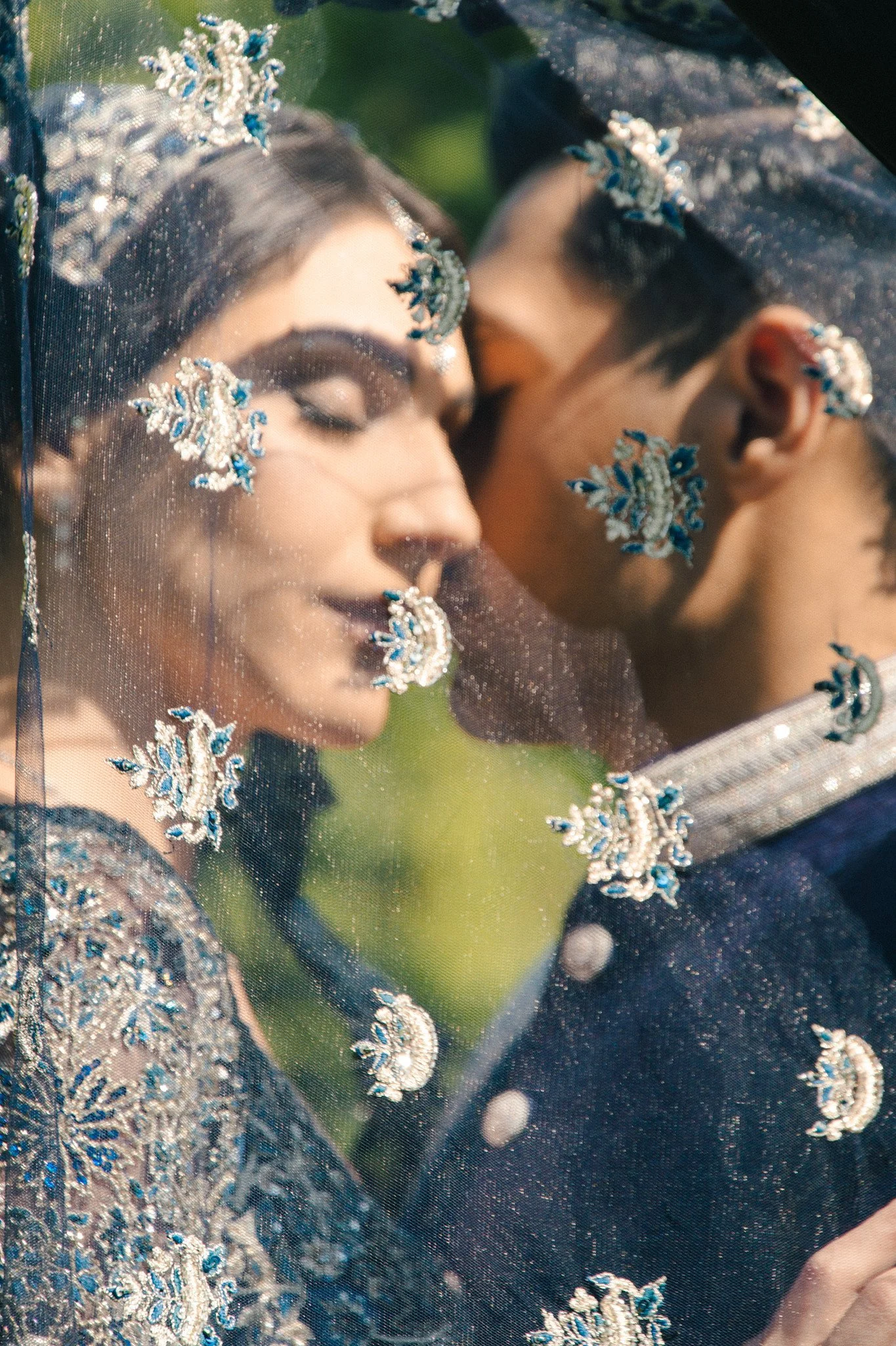 A bride and groom are seen through a decorative veil with blue and silver embellishments, sharing a close, intimate moment.