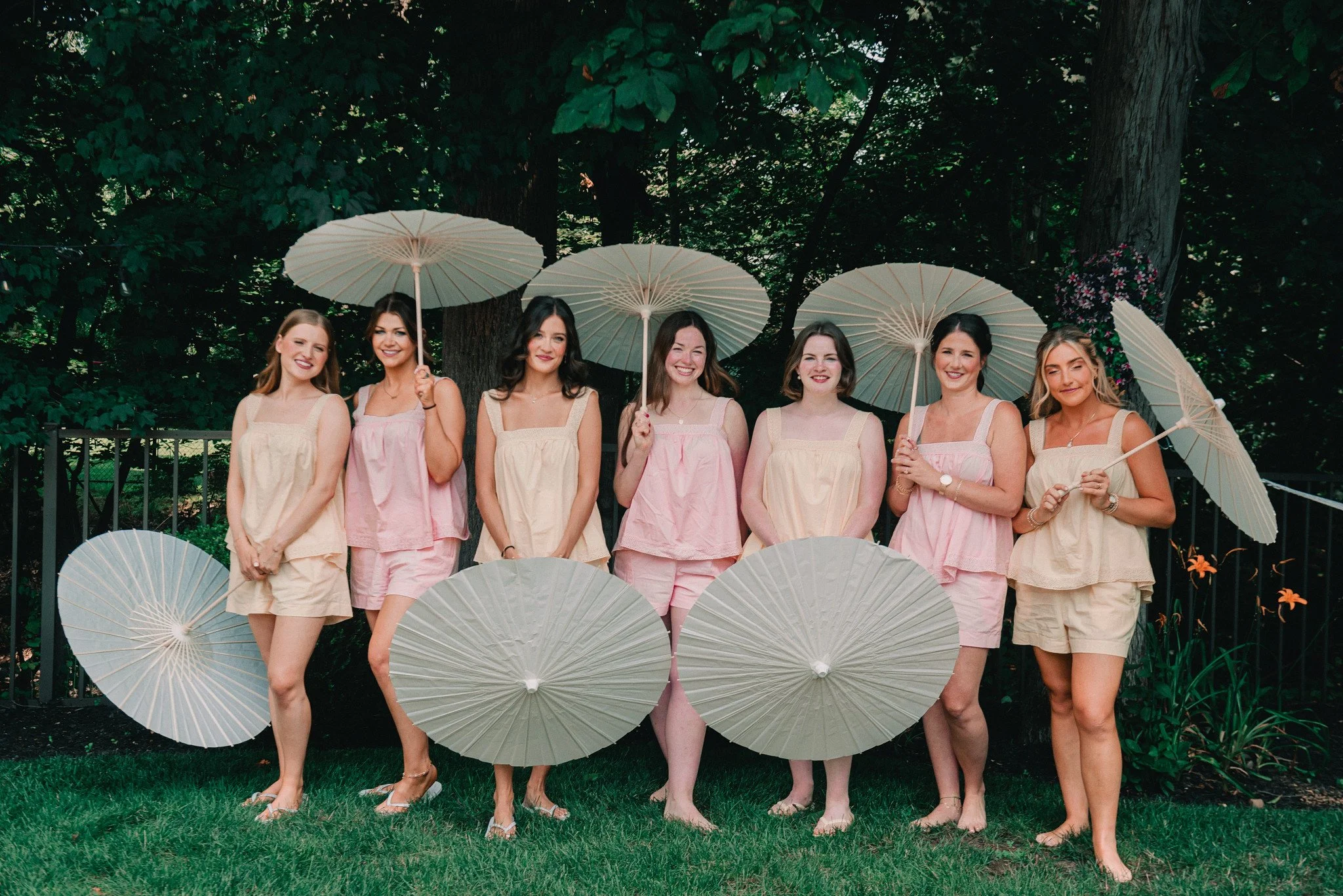 Seven women standing outdoors on grass, holding white parasols, wearing pastel-colored dresses, and smiling.
