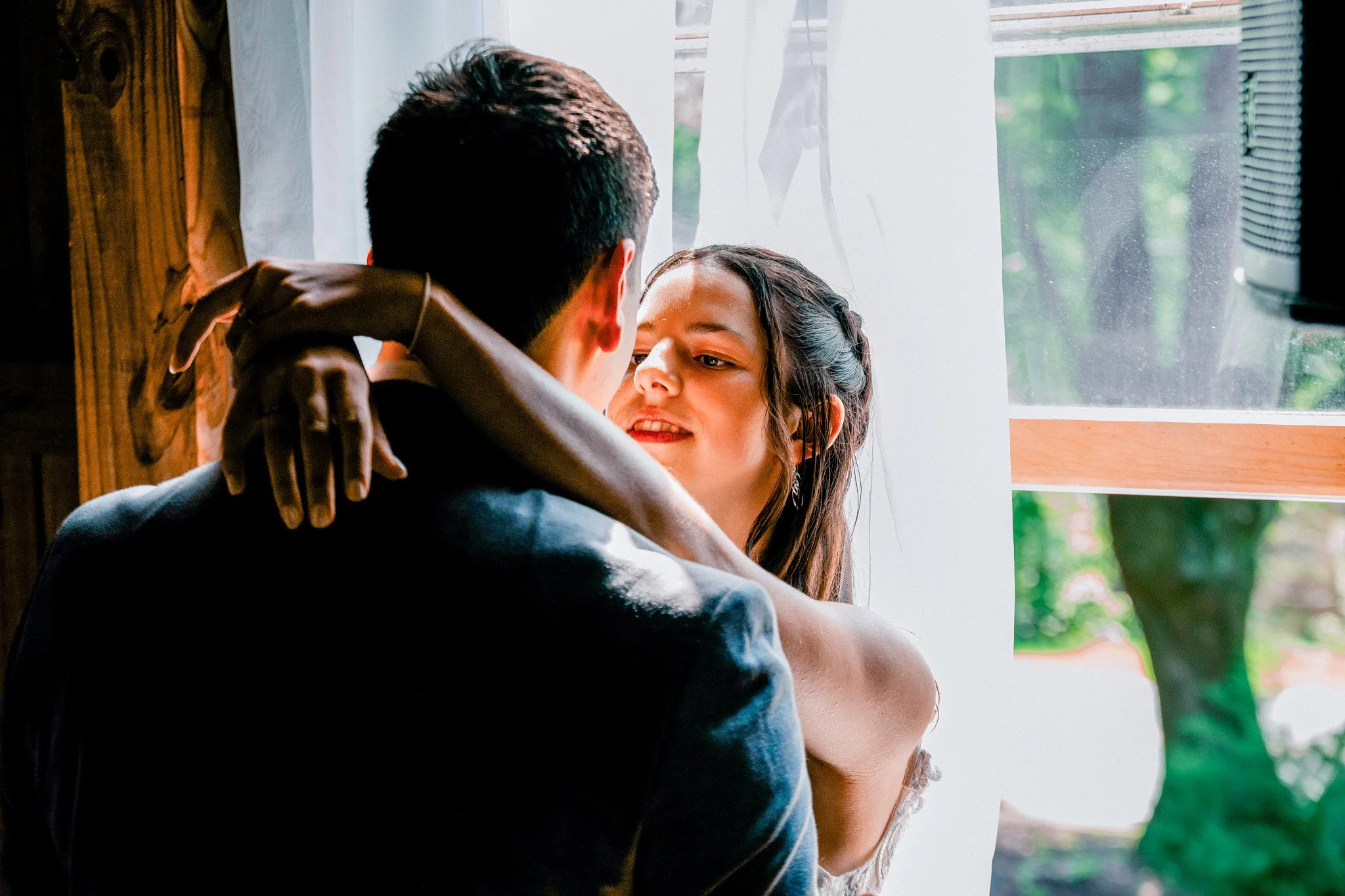 A man and woman embrace inside, near a window with sheer white curtains, with green trees visible outside.