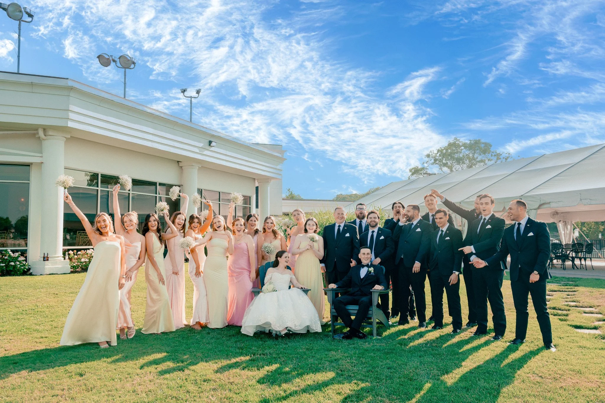 A wedding party posing on a sunny lawn outside a white building with a blue sky and scattered clouds in the background. The group includes bridesmaids in pastel dresses, groomsmen in suits, and a bride and groom sitting in front.