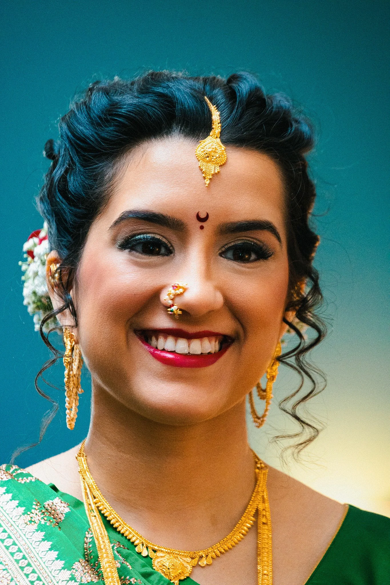 A smiling woman dressed in traditional Indian attire, wearing gold jewelry including earrings, necklaces, and a nose ring, with flowers in her braided hair and a red bindi on her forehead.