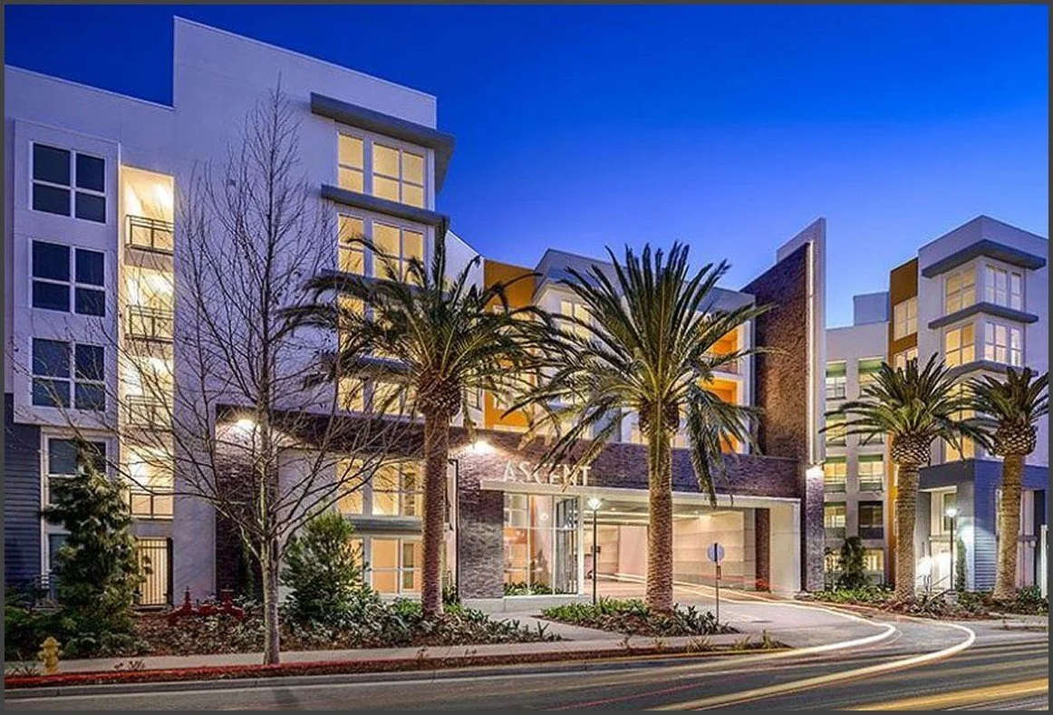 Modern multi-story apartment building illuminated at dusk with palm trees and landscaped area in front.