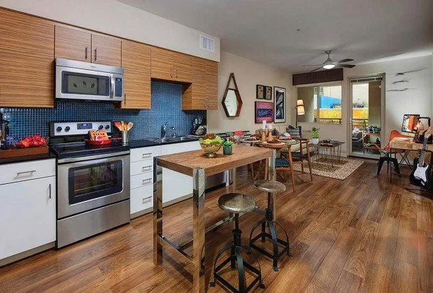 Open-concept living room and kitchen with wood flooring, modern kitchen appliances, a small kitchen island, and a cozy seating area with chairs, a desk, and large windows letting in natural light.