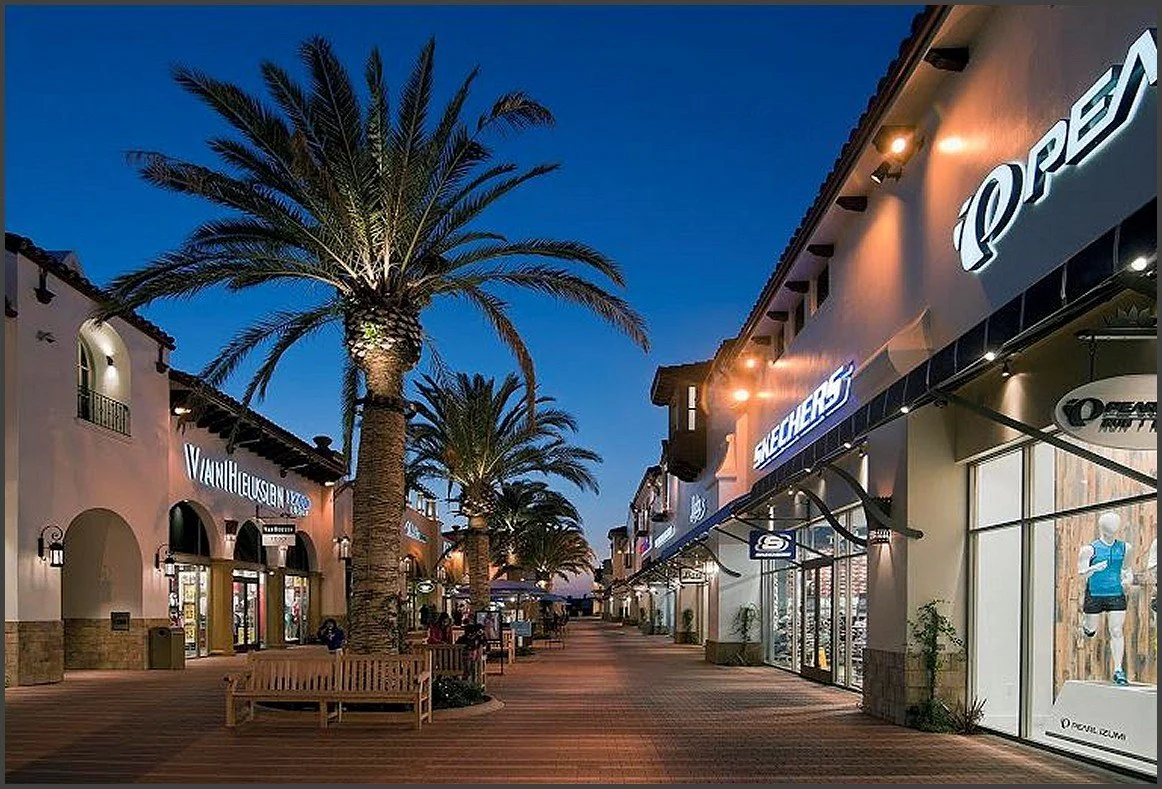 An outdoor shopping area with stores and palm trees at dusk.