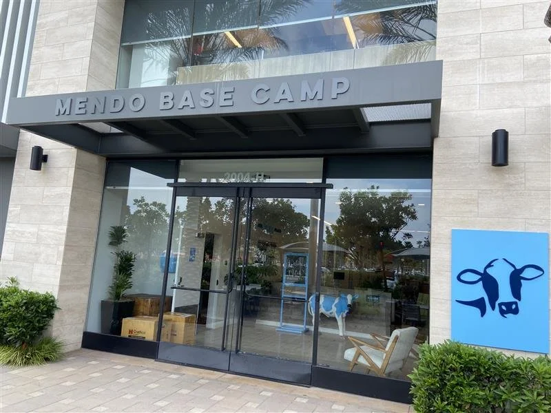 The entrance to Mendo Base Camp with glass doors, a cow logo on a blue sign, and a cow statue inside visible through the glass.