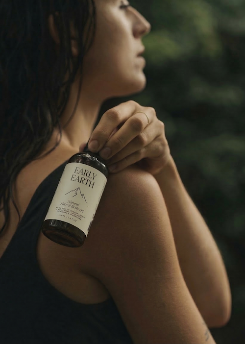 A woman with dark, wet hair is holding a bottle of Early Earth natural herbal oil against her shoulder outdoors, with a blurred green background.