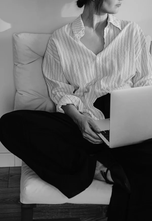 A woman sitting on a couch using a laptop, wearing a striped button-up shirt and black pants, in a minimalistic room.
