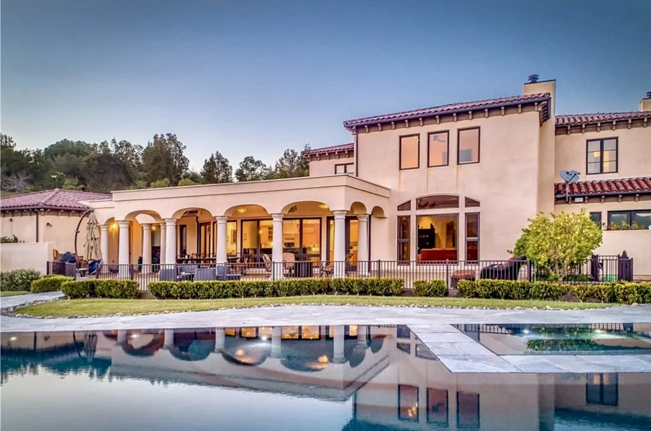 Modern house with a swimming pool in the foreground, illuminated outdoor patio with columns, trees, and a hillside in the background at dusk.