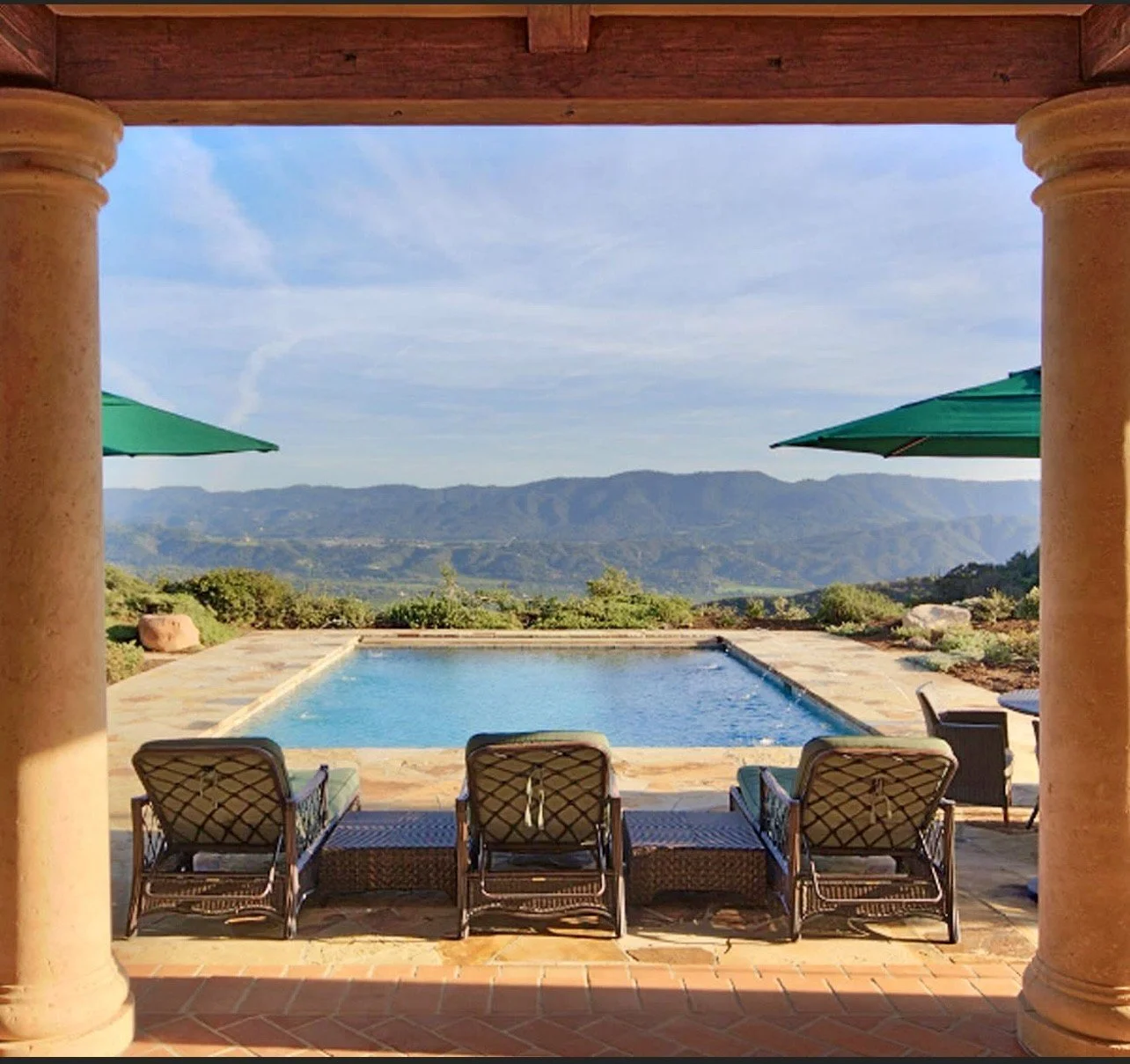 View of a swimming pool with outdoor chairs and umbrellas, overlooking a landscape of rolling hills and mountains.