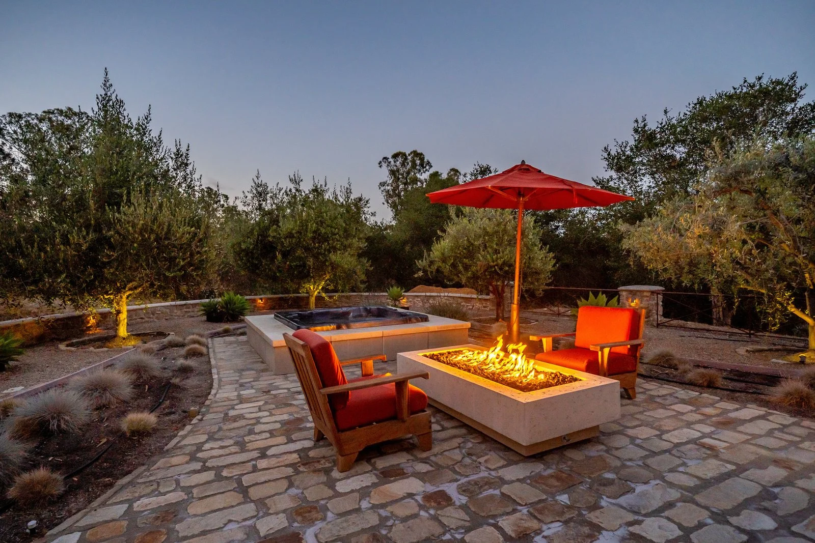 Outdoor patio with a fire pit, two wooden armchairs with red cushions, a red umbrella, and a hot tub, surrounded by trees and plants at dusk.