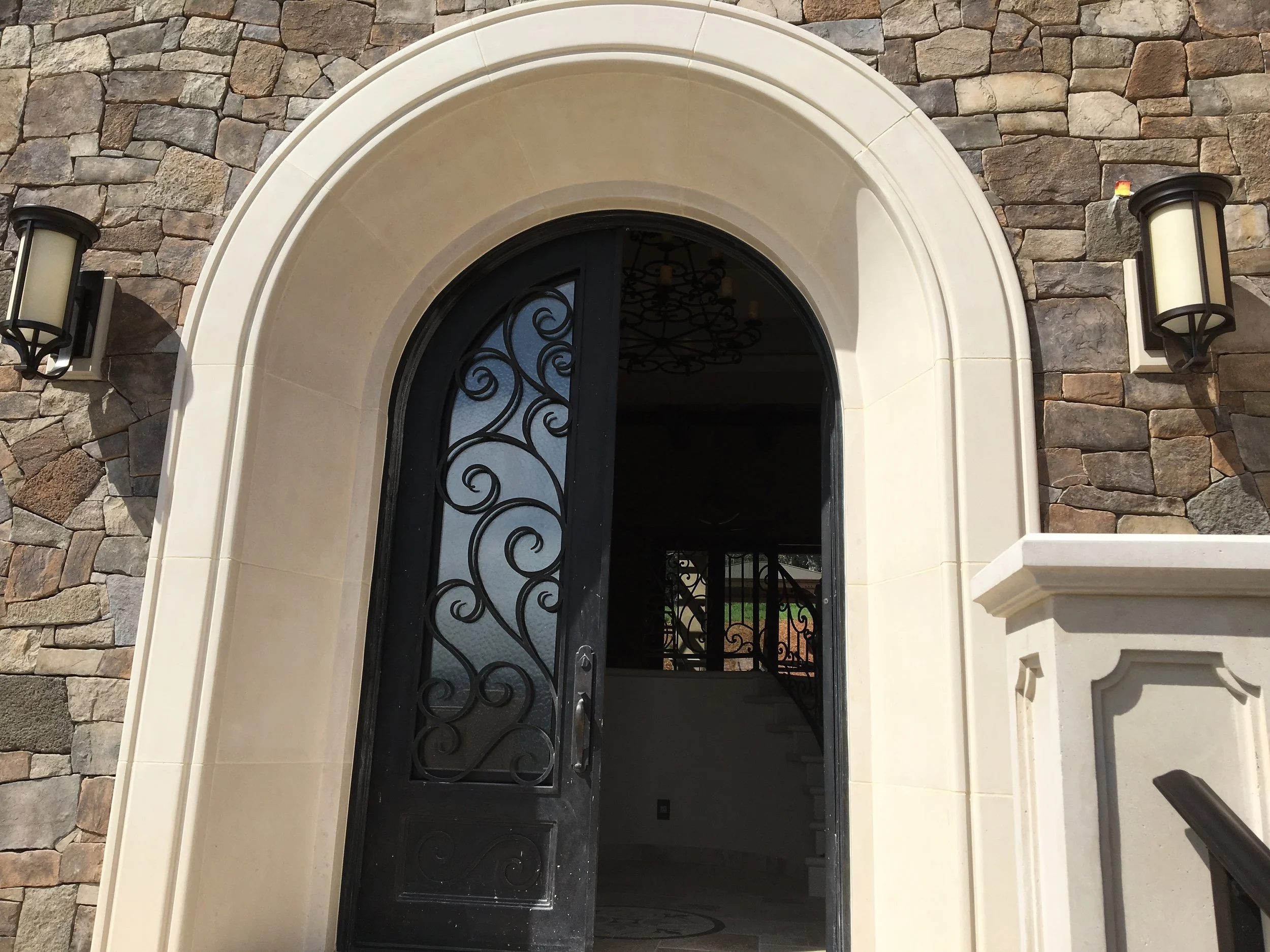 A decorative arched entrance with a black wrought iron door featuring intricate scrollwork. The door is framed by cream-colored stone trim, flanked by two stone walls made of irregularly shaped stones. Two wall-mounted lanterns illuminate the entranc