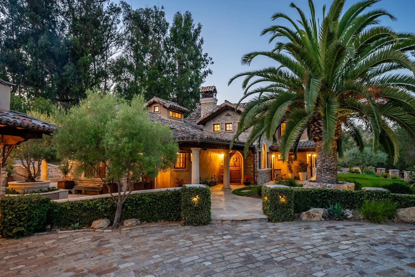 A house with a stone exterior, warm lit windows, surrounded by palm trees and greenery on a cobblestone driveway at dusk.