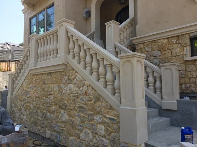 Front view of a stone and stucco house with a staircase leading to the front entrance, featuring decorative balusters and columns.