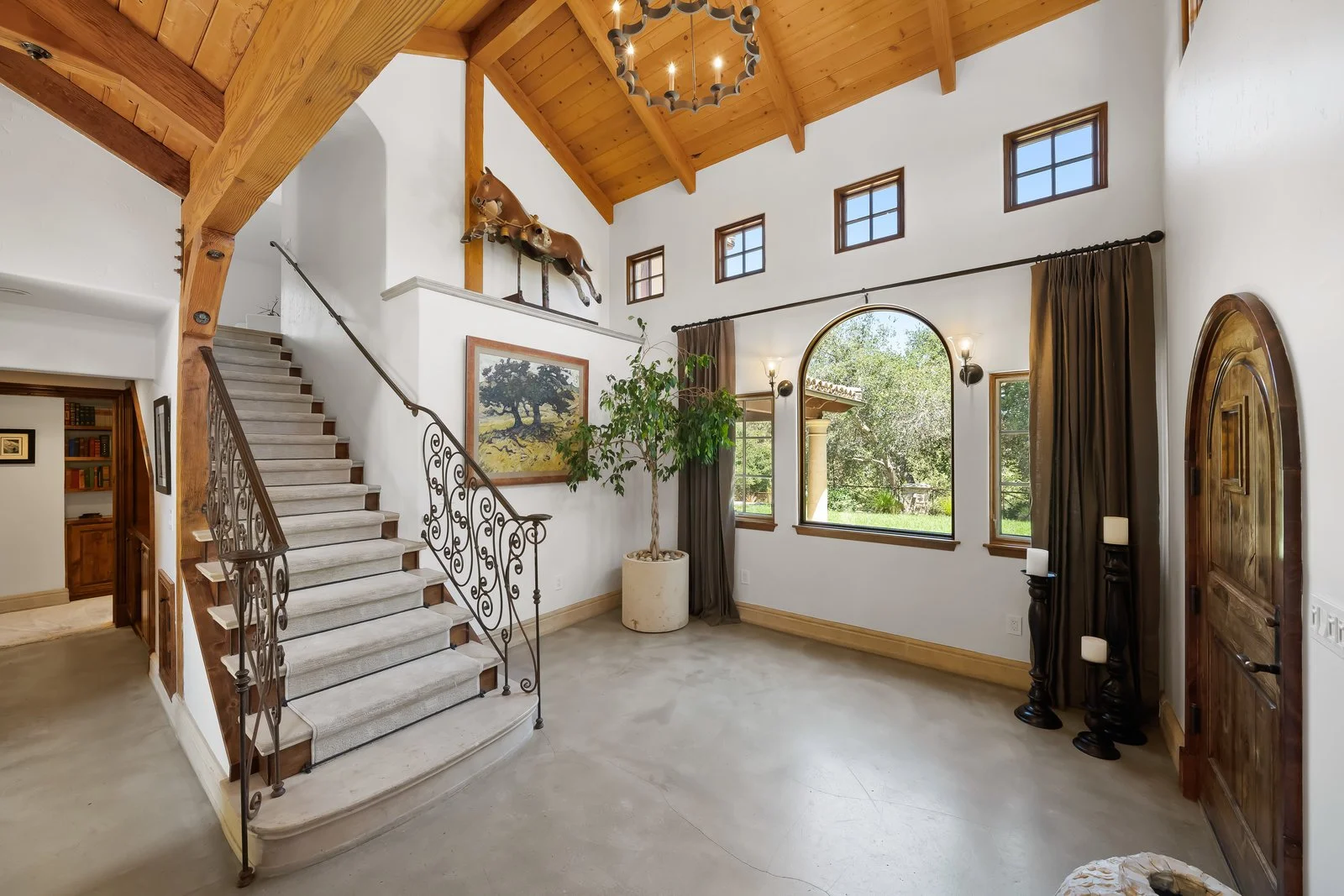 Interior view of a spacious living room with a high wooden ceiling, large windows, a staircase with decorative wrought-iron railings, potted plant, artwork, and a wooden door.